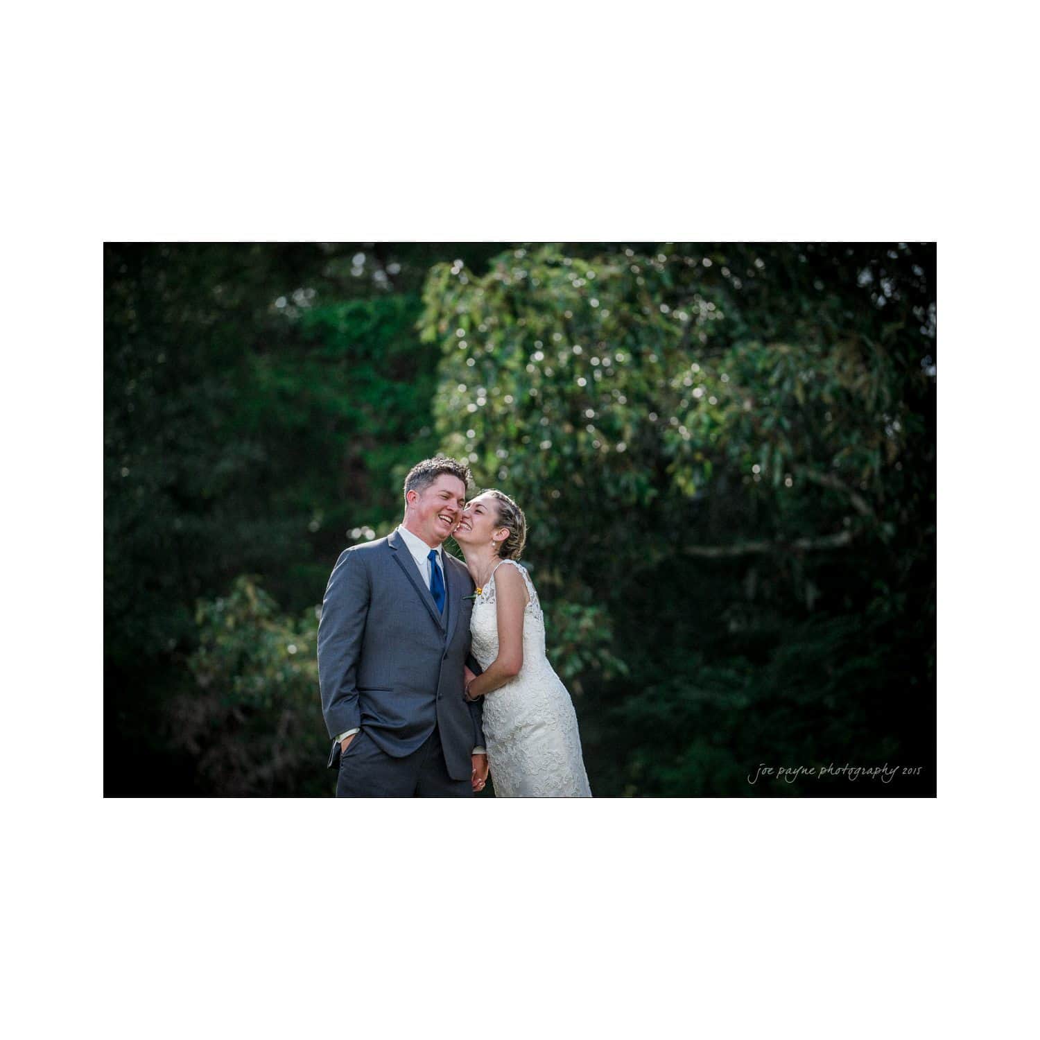 chapel hill wedding photography - nicole & wayne A bride in a lace dress and a groom in a gray suit stand smiling outdoors, with the bride leaning in affectionately toward the groom. They are surrounded by lush greenery.