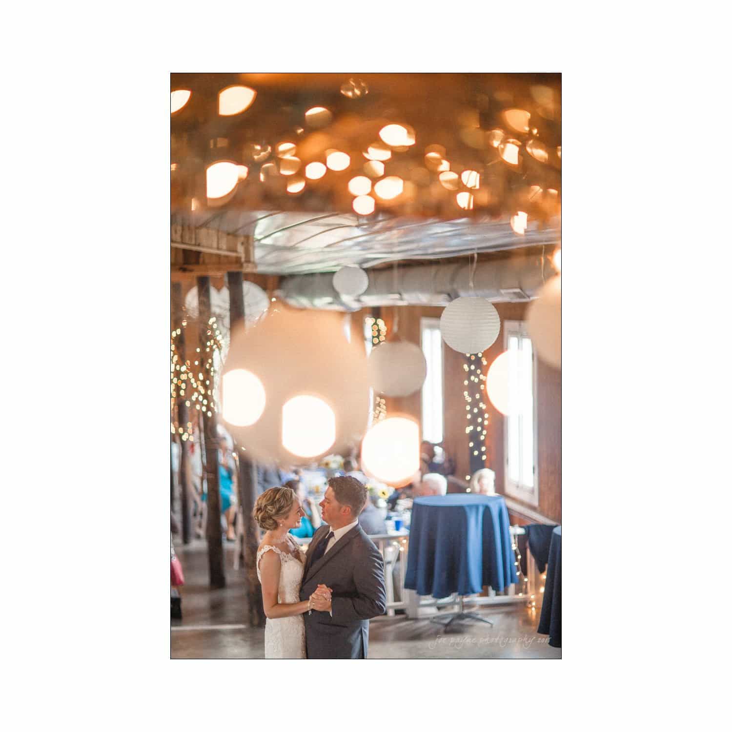 chapel hill wedding photography - nicole & wayne A couple dances closely in a warmly lit venue, decorated with hanging paper lanterns and string lights. Large windows are visible in the background, with guests seated at tables covered in blue tablecloths.