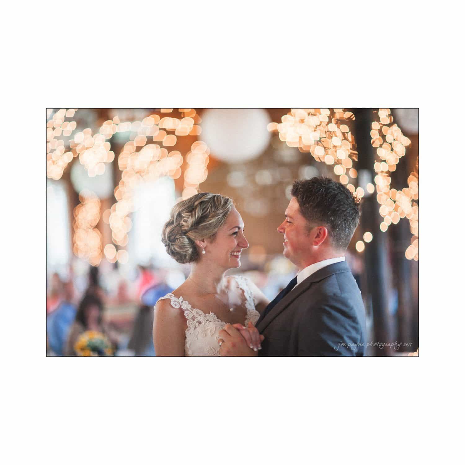 chapel hill wedding photography - nicole & wayne A couple shares a joyful dance at their wedding, surrounded by a soft, blurred background of warm lights and decorations. They are smiling at each other, creating a romantic and festive atmosphere.