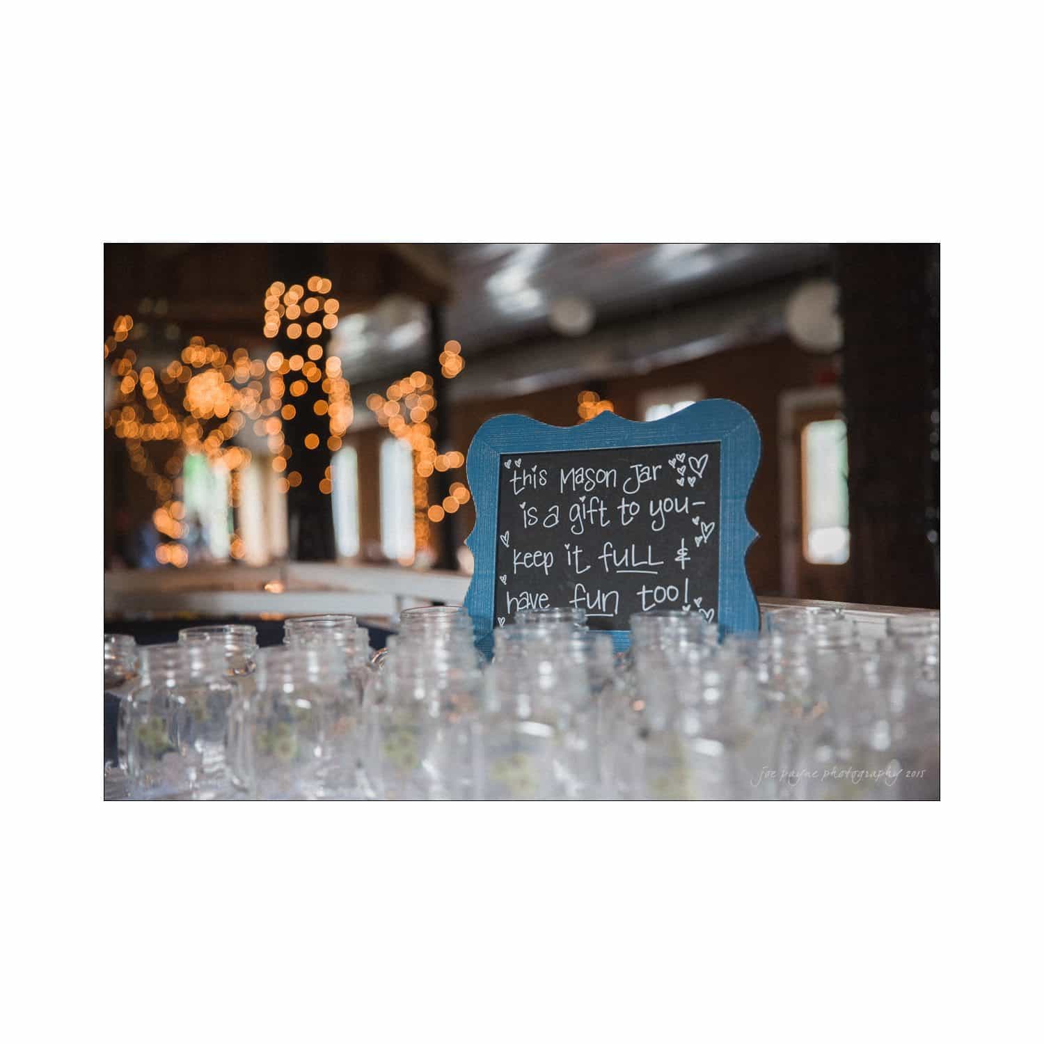 chapel hill wedding photography - nicole & wayne A table with empty mason jars and a chalkboard sign in a dimly lit room. The sign reads, This mason jar is a gift to you - keep it full & have fun too! Twinkling string lights are visible in the background.