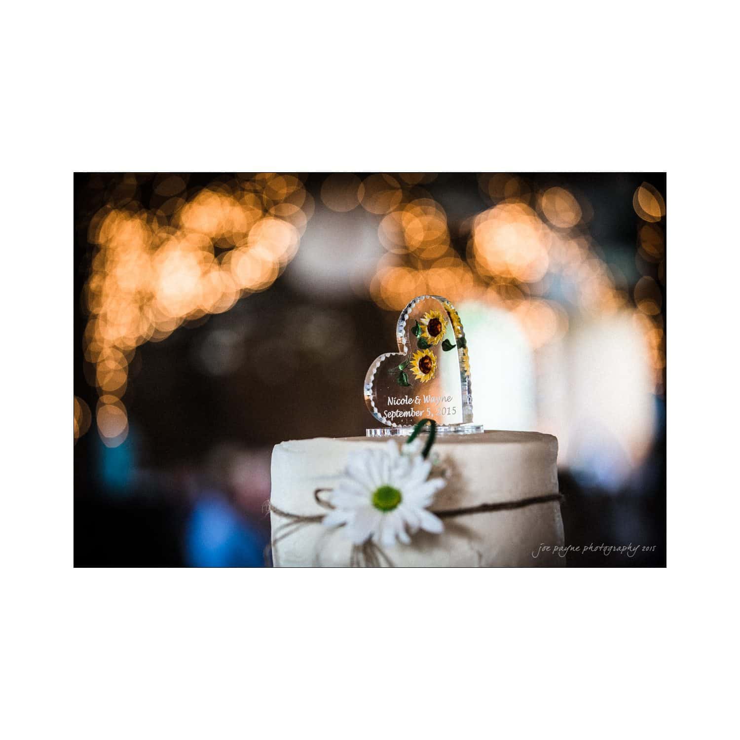 chapel hill wedding photography - nicole & wayne Close-up of a heart-shaped cake topper with sunflowers and the text, Nicole & Mike, September 3, 2015, on a tiered cake adorned with a daisy. The background features blurred, warm string lights.