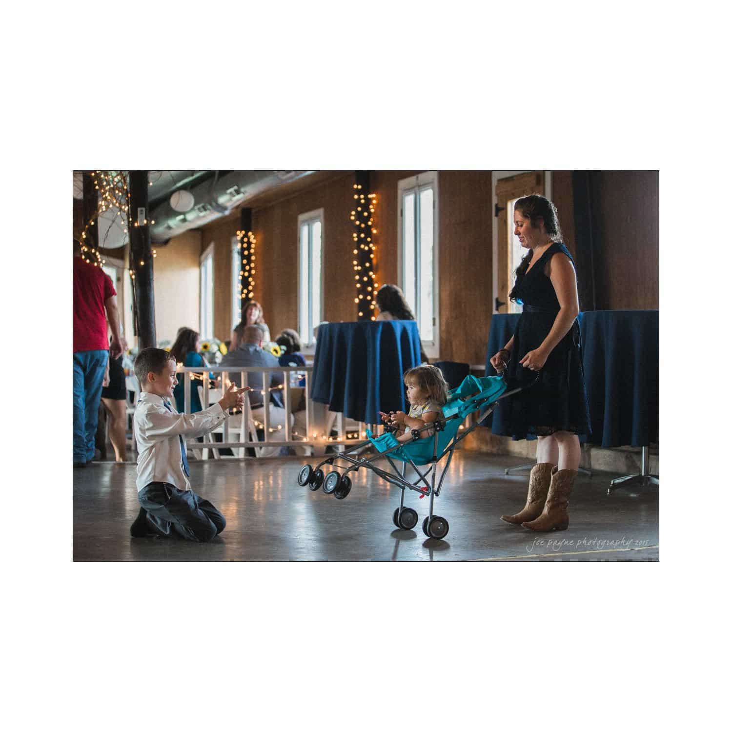 chapel hill wedding photography - nicole & wayne A young woman in a black dress and boots pushes a baby in a stroller. A boy kneels in front, interacting playfully. They are in a decorated room with string lights and tables. Guests in the background enjoy the event.
