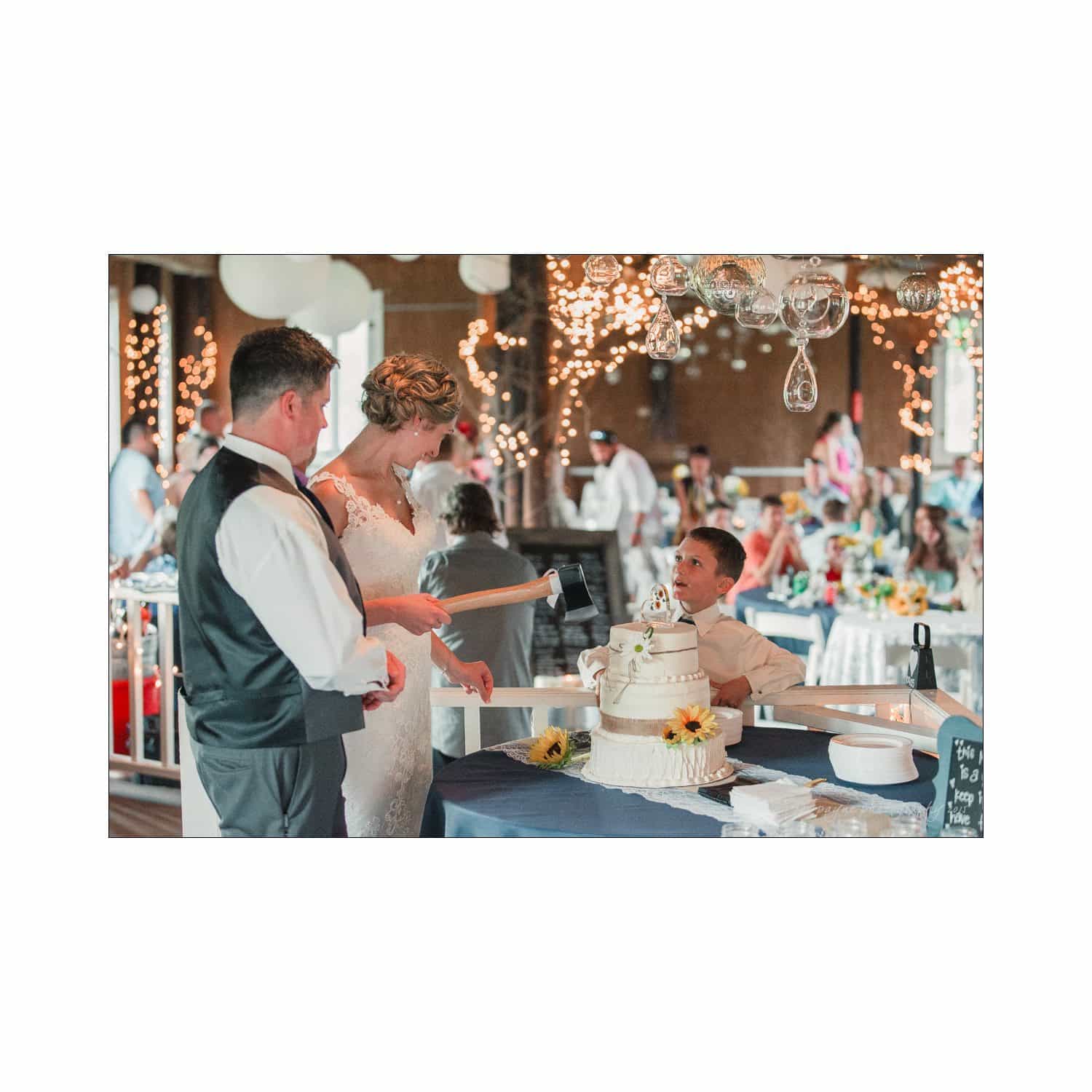 chapel hill wedding photography - nicole & wayne A bride and groom stand beside a three-tier wedding cake adorned with sunflowers. A young boy in a white shirt assists them in cutting the cake. The background features guests and ornate string lights in a warmly lit venue.