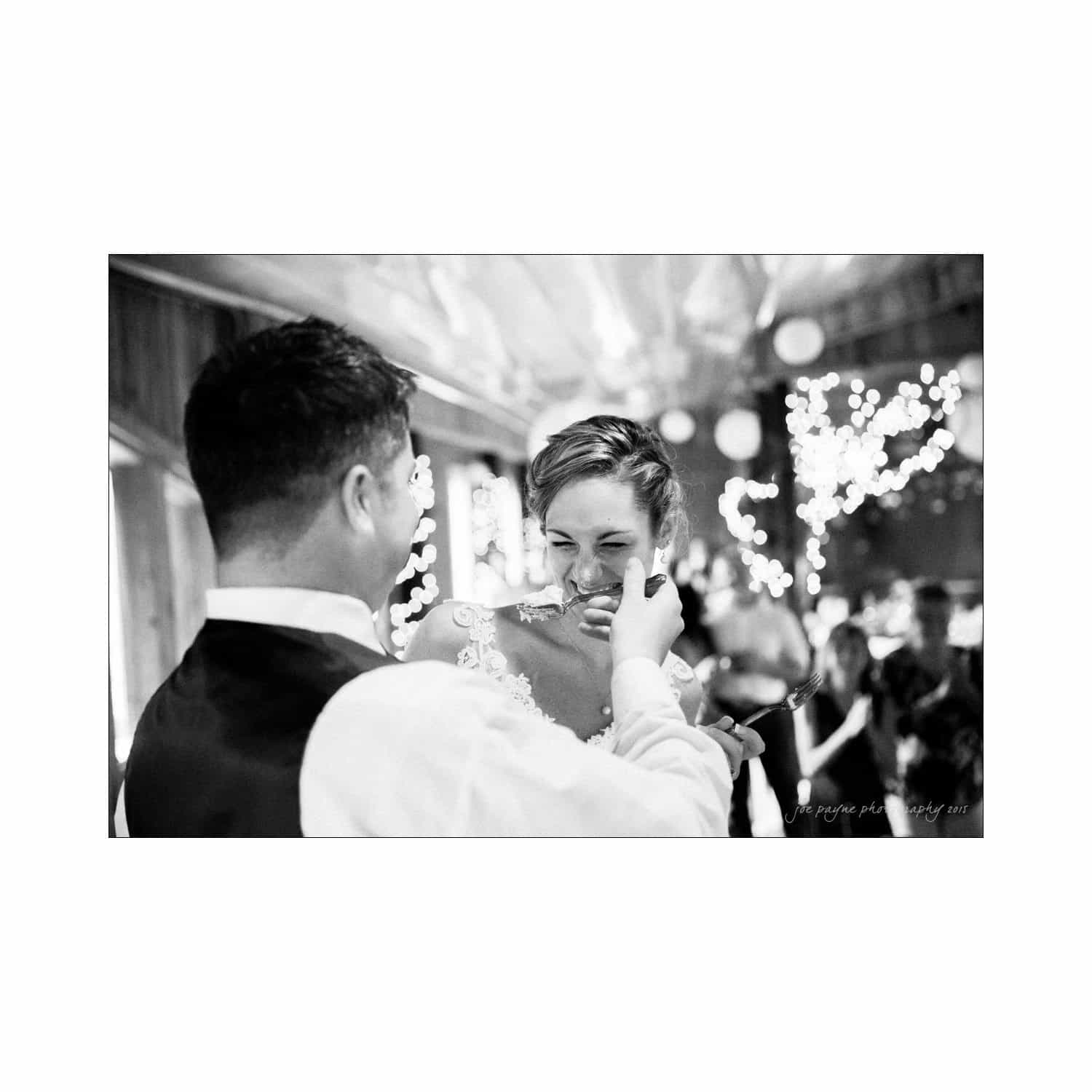 chapel hill wedding photography - nicole & wayne A bride and groom joyfully feed each other cake at a wedding reception. The scene is set indoors with soft, ambient lighting and blurred guests in the background, capturing a moment of celebration and laughter.