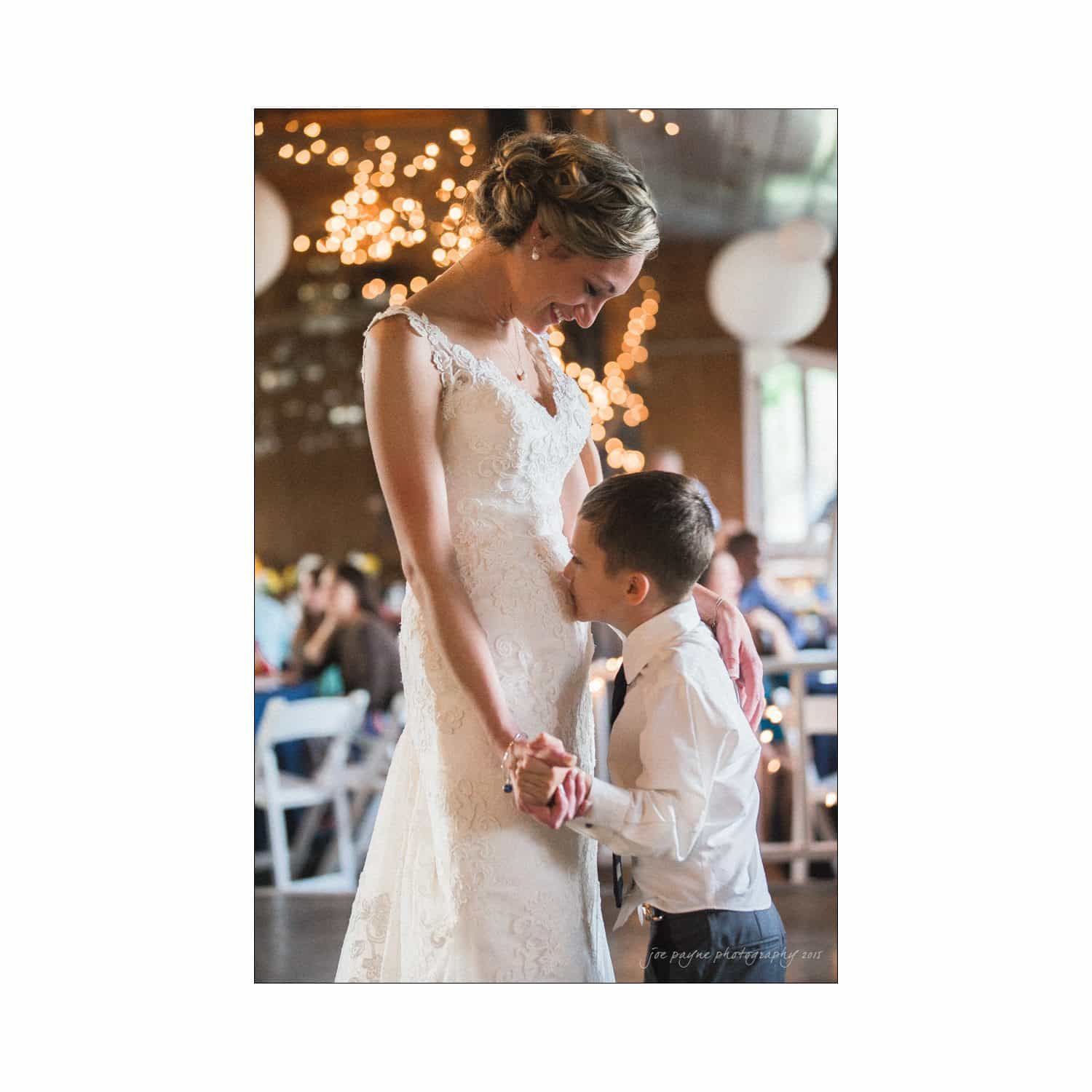 chapel hill wedding photography - nicole & wayne A bride in a white lace dress smiles warmly as a young boy, dressed in a white shirt and tie, hugs her tightly during a dance. Soft, warm lights create a cozy atmosphere in a festive setting.