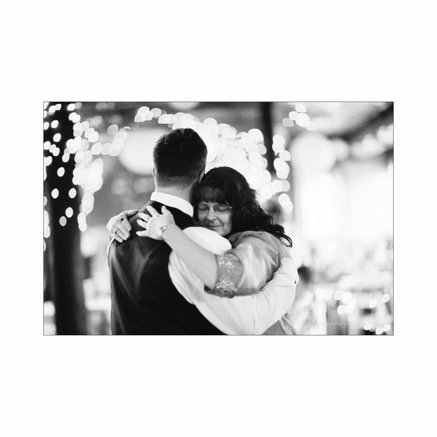 chapel hill wedding photography - nicole & wayne A black and white photo of a joyful embrace between two people, surrounded by soft, twinkling lights in the background. The setting suggests a celebratory or formal event.