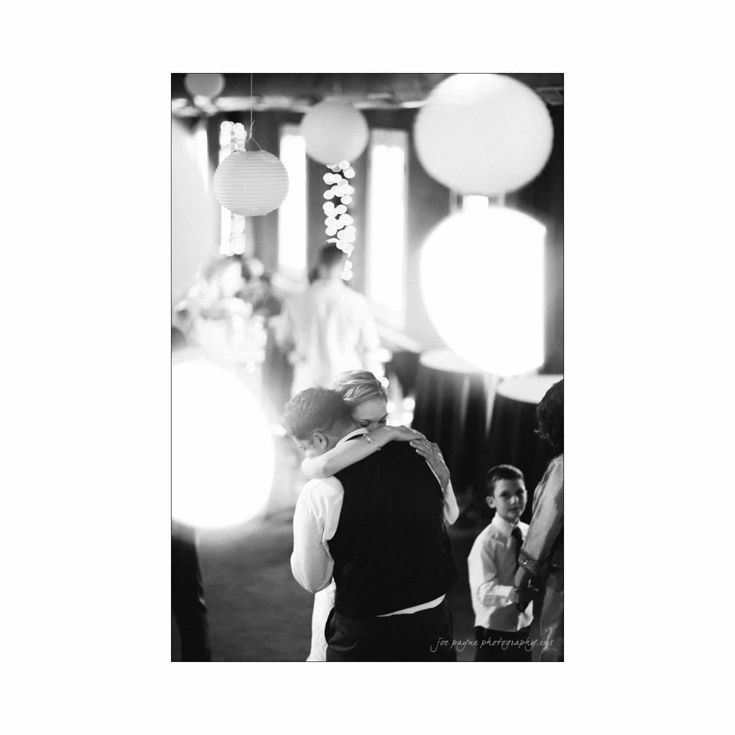 chapel hill wedding photography - nicole & wayne A black and white photo of a couple embracing on a dance floor, surrounded by large hanging lanterns. Guests are visible in the background, seated and standing. The ambiance is intimate and celebratory.