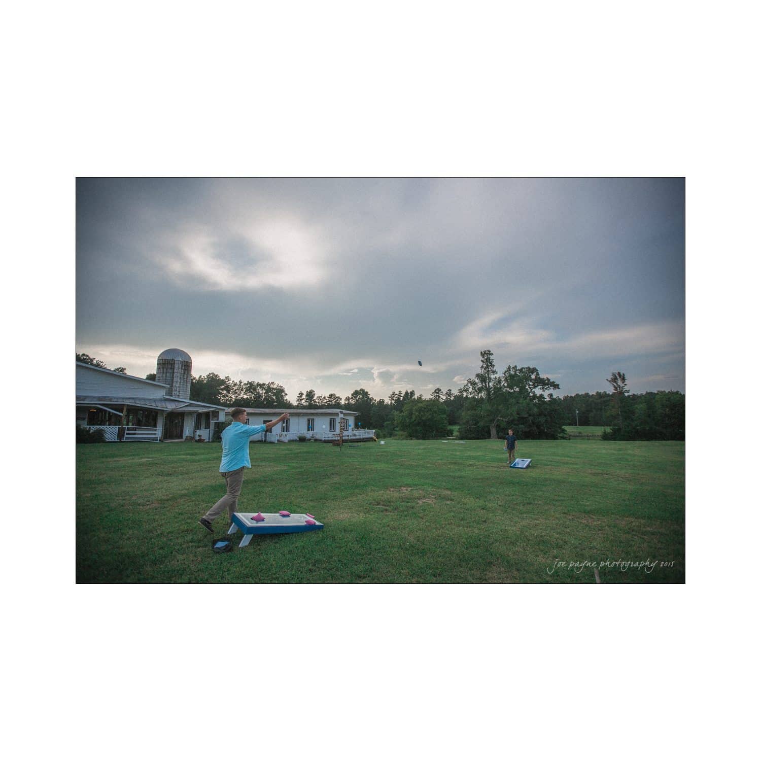 chapel hill wedding photography - nicole & wayne Two people playing cornhole on a grassy field. One person throws a bean bag towards a board, while the other stands near a second board. A house and trees are in the background under a cloudy sky.