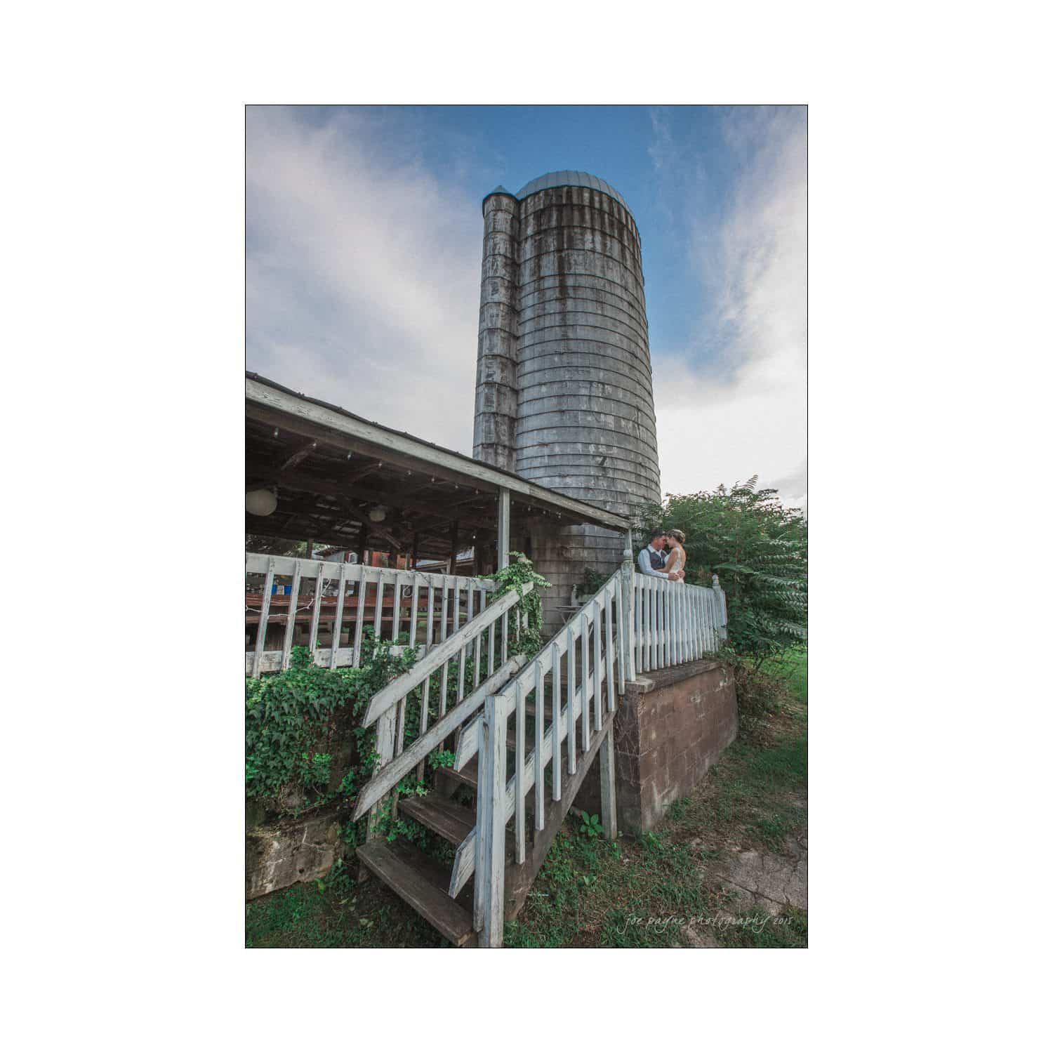 chapel hill wedding photography - nicole & wayne A rustic farm scene featuring a large, weathered silo next to a wooden building with a white railing and stairs. Green foliage surrounds the structure, and the sky is partly cloudy, creating a peaceful, rural atmosphere.