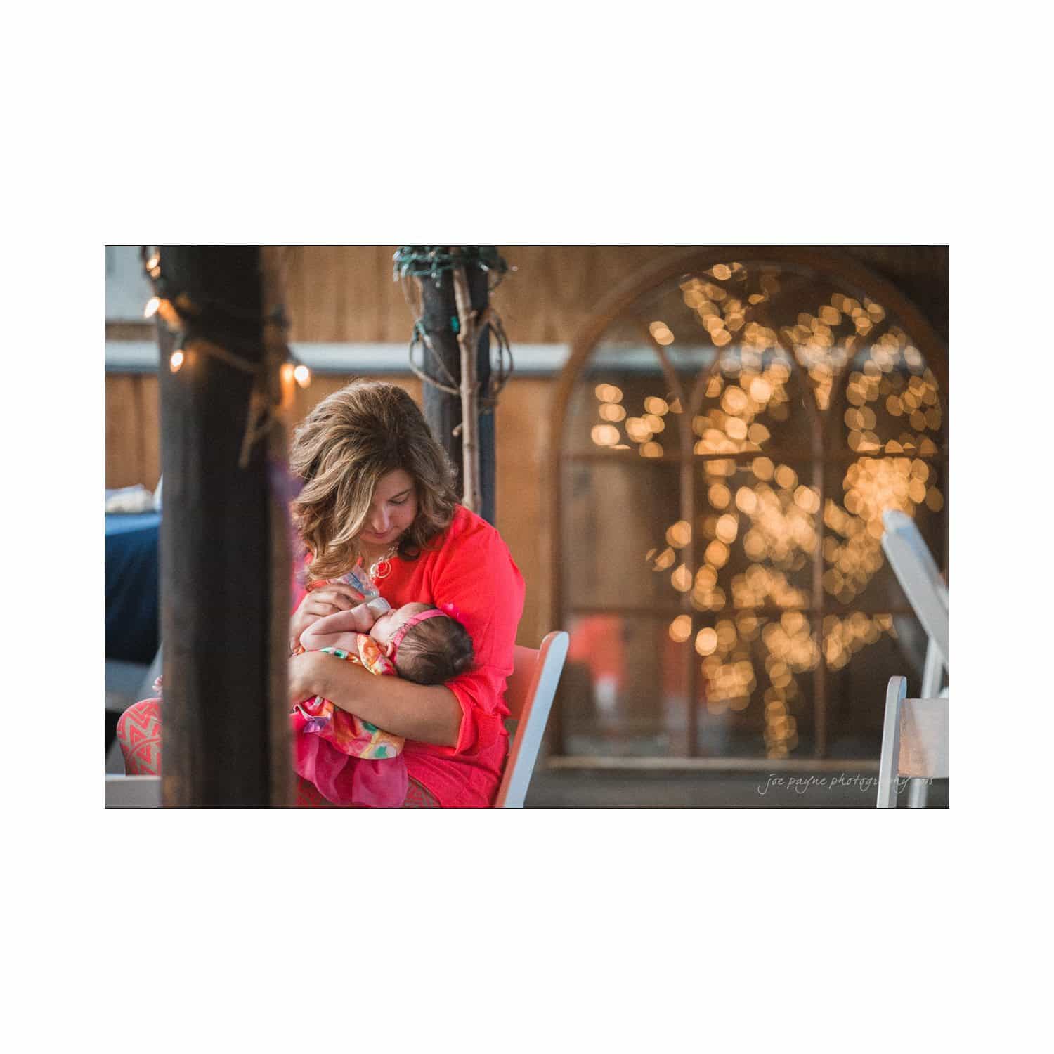 chapel hill wedding photography - nicole & wayne A woman in a pink top sits on a chair, gently holding and looking at a baby. Warm fairy lights create a soft, bokeh effect in the background, enhancing the cozy atmosphere.
