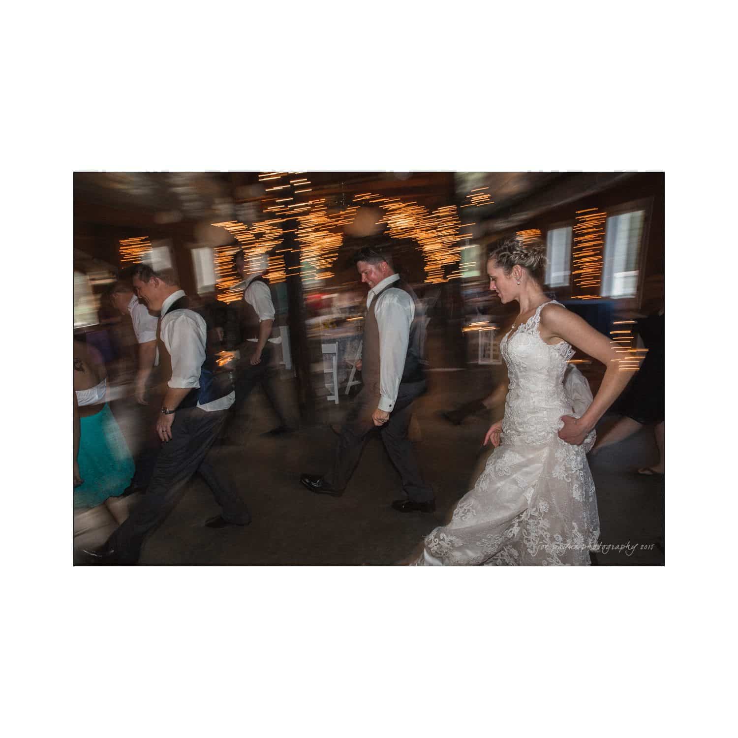 chapel hill wedding photography - nicole & wayne A bride in a white dress joins a line dance at a wedding reception. She is surrounded by other guests, all moving in rhythm. Twinkling lights decorate the background, creating a festive atmosphere.