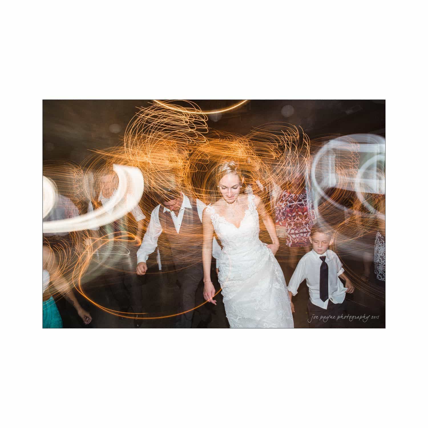 chapel hill wedding photography - nicole & wayne A bride in a white dress dances energetically with a young boy and others. The scene is vibrant, with swirling lights creating dynamic patterns in the background, capturing the joyous atmosphere of the celebration.