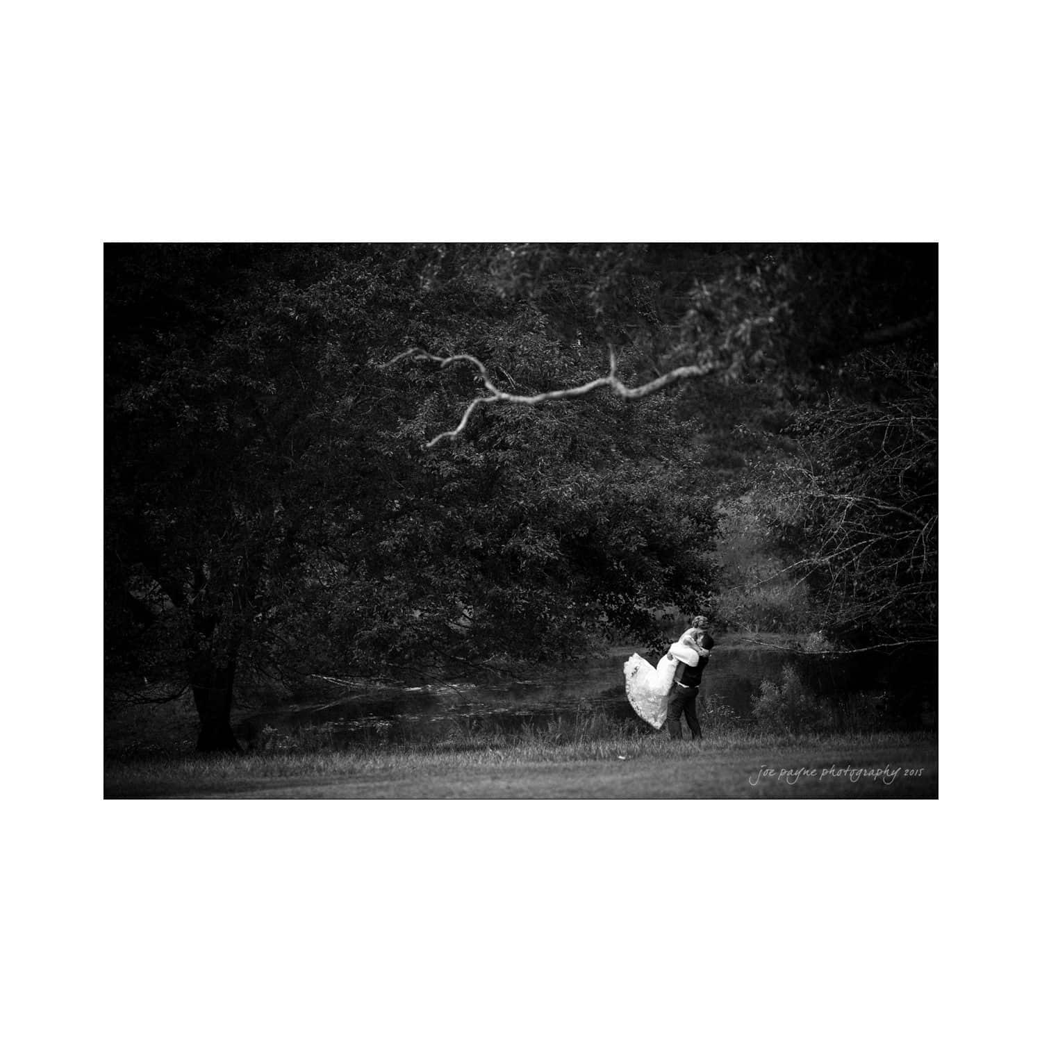 chapel hill wedding photography - nicole & wayne A couple in wedding attire embrace and kiss under a large tree in a park. The image is in black and white, highlighting the romantic and serene atmosphere.