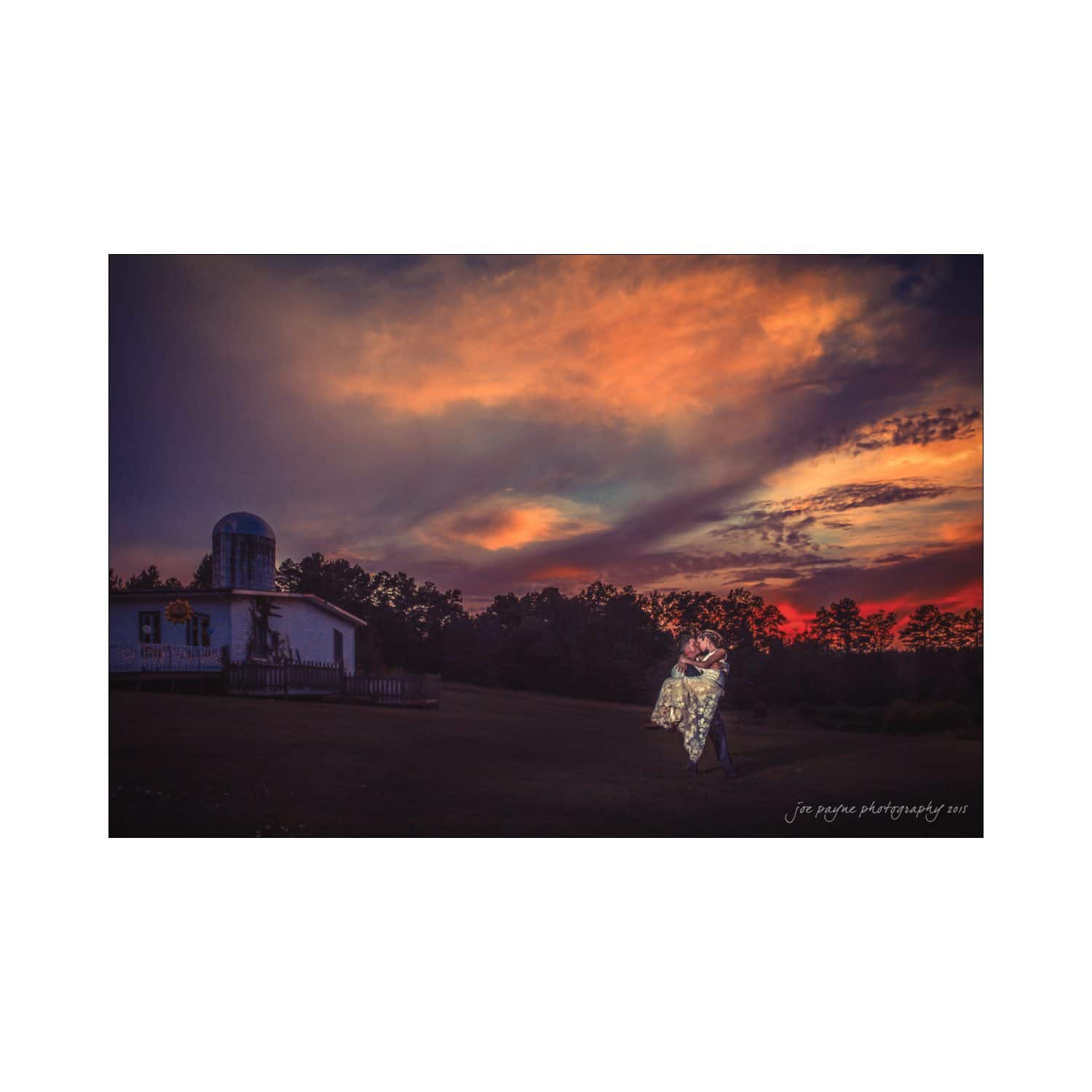 chapel hill wedding photography - nicole & wayne A couple embraces under a dramatic, colorful sunset sky. A small white building with a dome and trees are visible in the background, creating a serene, romantic scene.