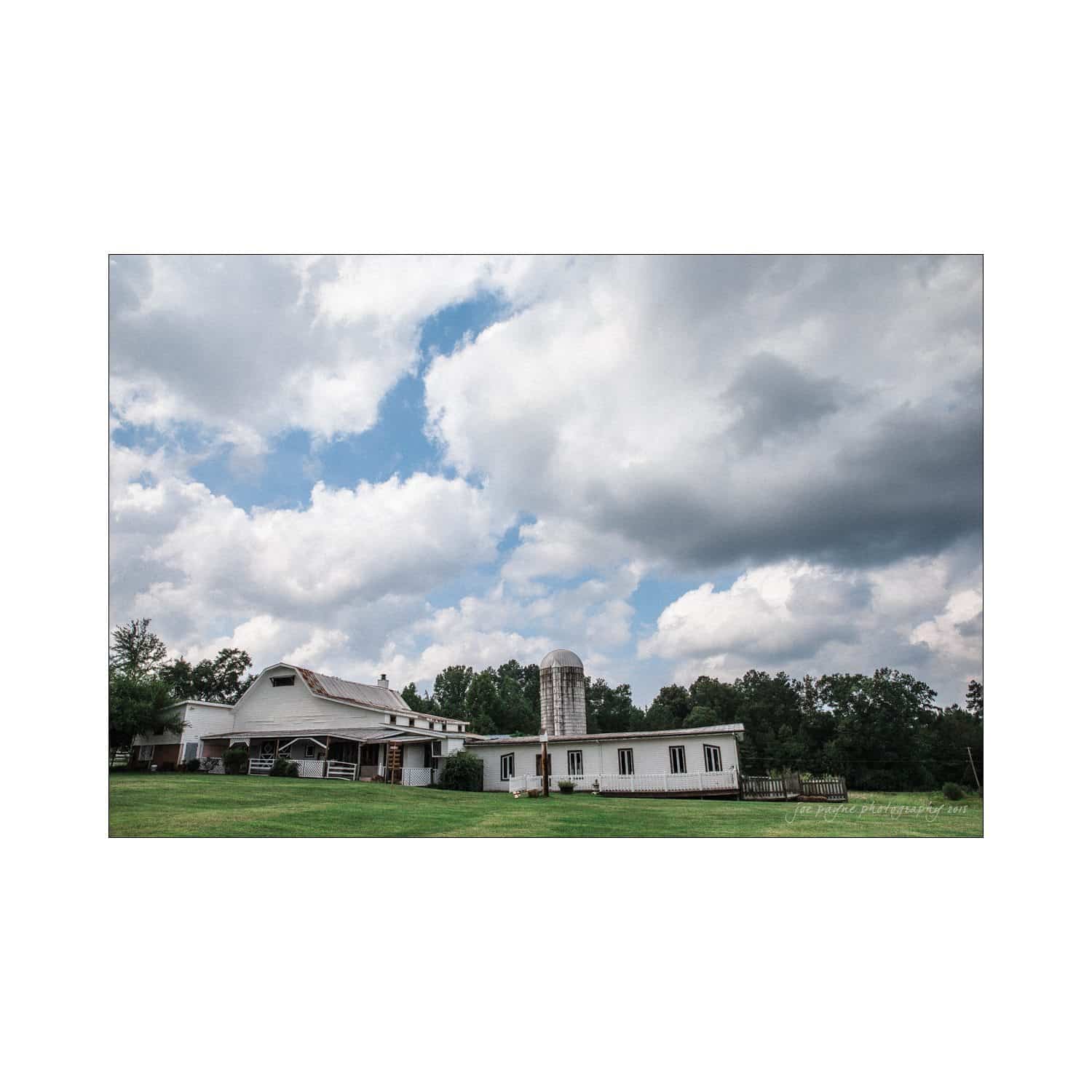 chapel hill wedding photography - nicole & wayne A scenic view of a white barn and silo under a partly cloudy sky. The barn is surrounded by lush green grass and trees. The sky is a mix of white fluffy clouds and patches of blue.