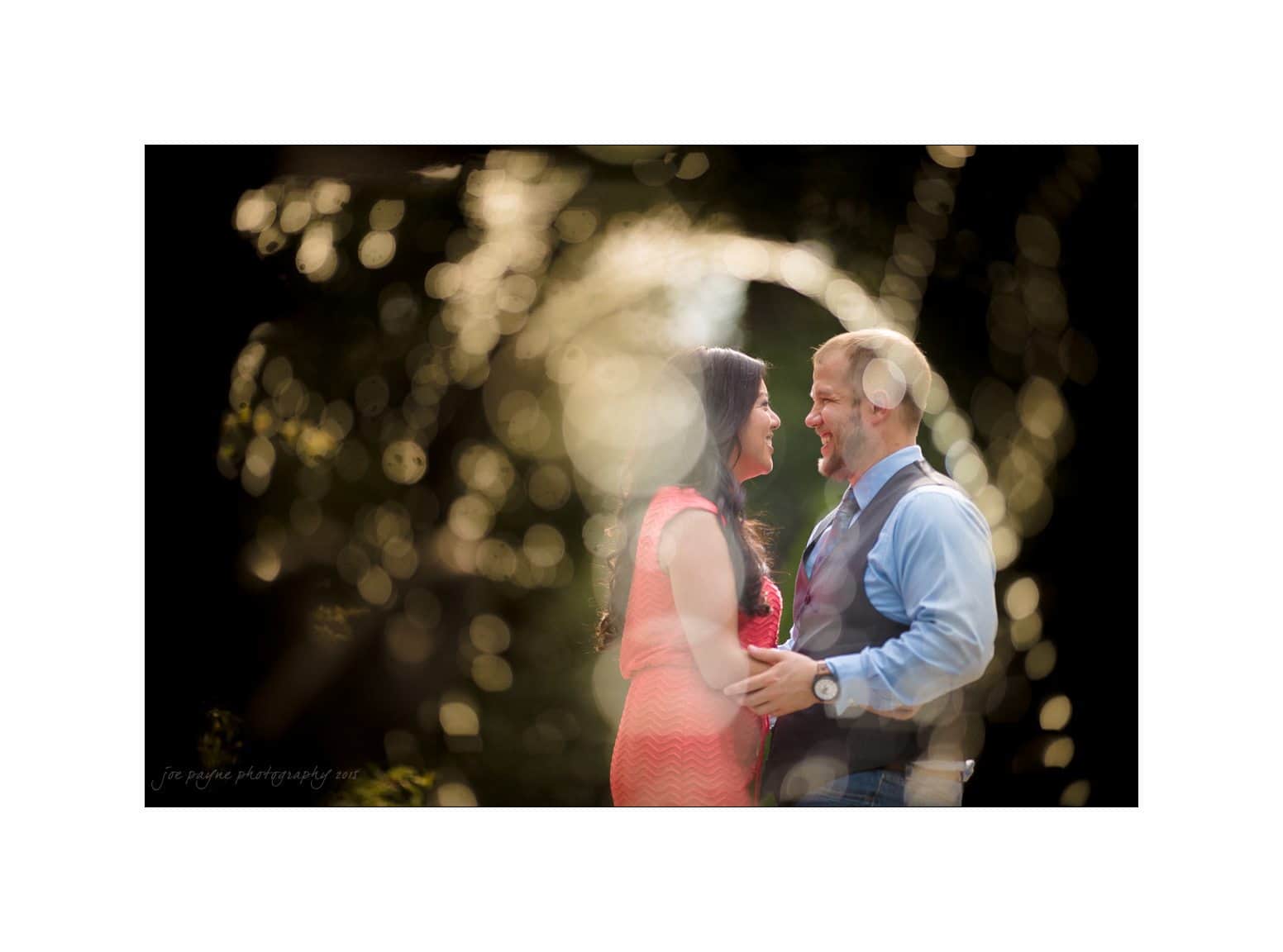 A couple stands facing each other outdoors, holding hands and smiling. The background is artistically blurred with circular light effects, creating a dreamy atmosphere. The woman is in a pink dress, and the man is in a blue shirt and vest.