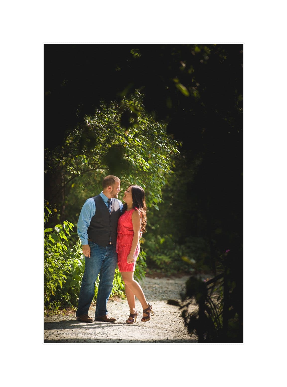 A couple stands on a sunlit path surrounded by greenery. The man is wearing jeans and a blue shirt with a vest, and the woman is in a red dress. They are looking at each other, smiling, and holding hands.