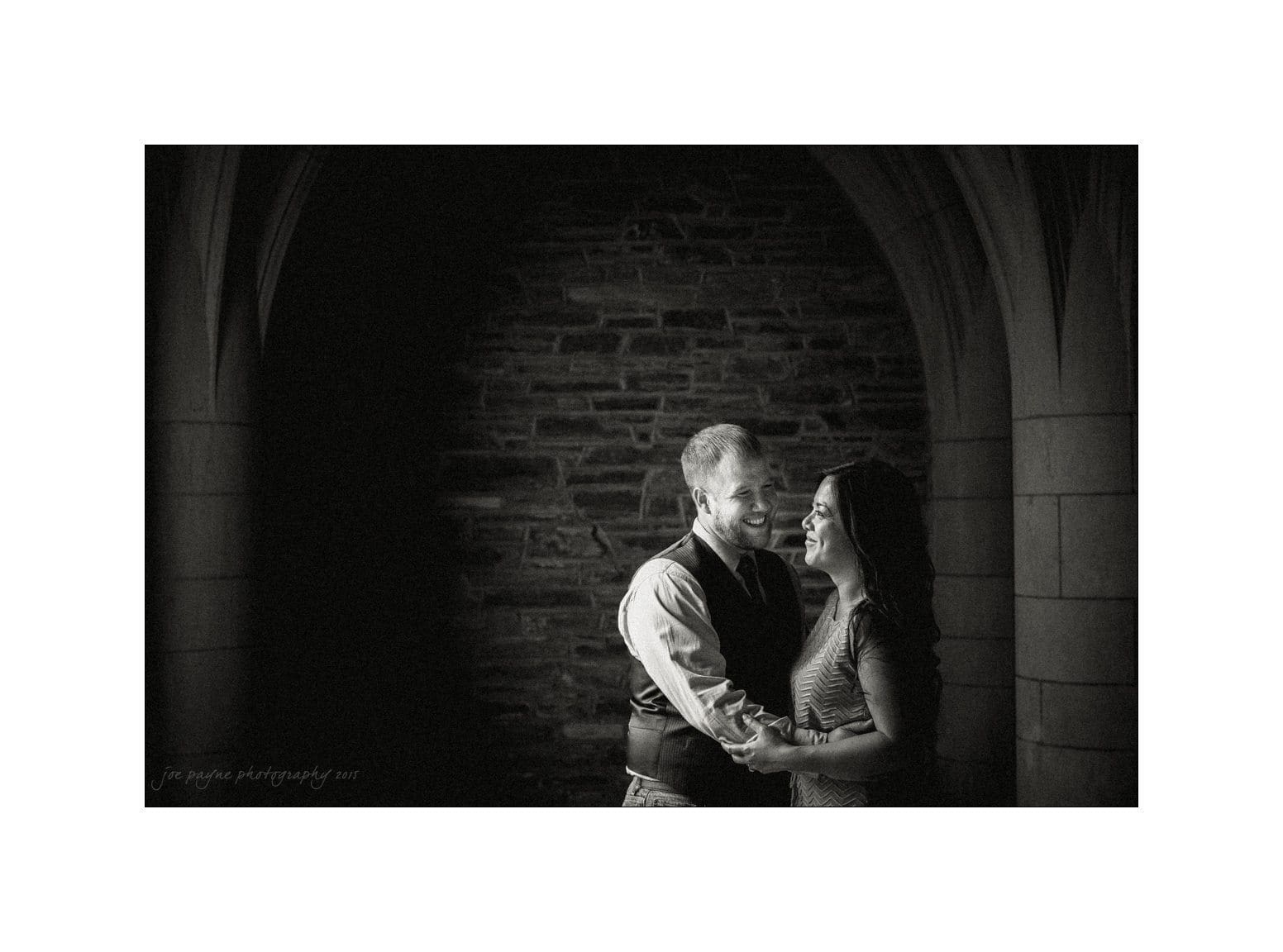 A couple stands close together, smiling and holding hands, in an arched stone doorway. The background is dark, contrasting with the couples joyful expressions, creating an intimate and romantic atmosphere.