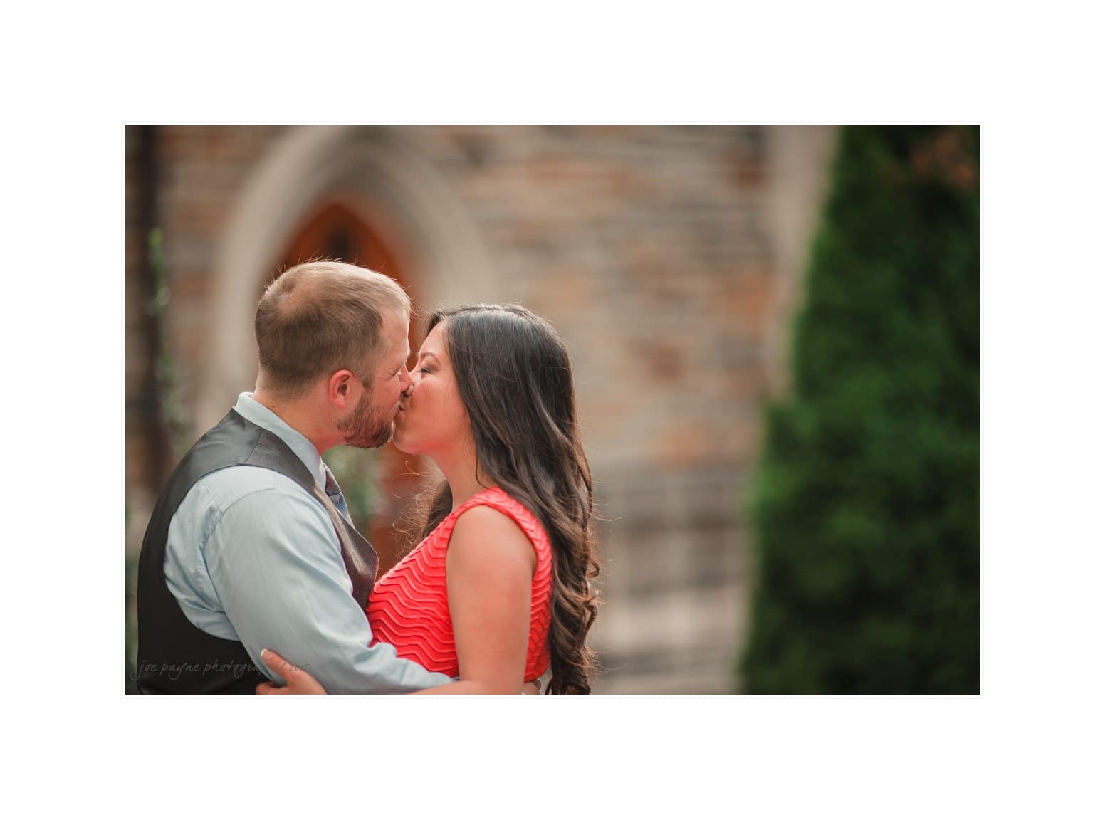A couple shares a kiss in front of a stone building with arched windows. The man wears a blue shirt and gray vest, while the woman wears a red dress. Green foliage can be seen in the background.
