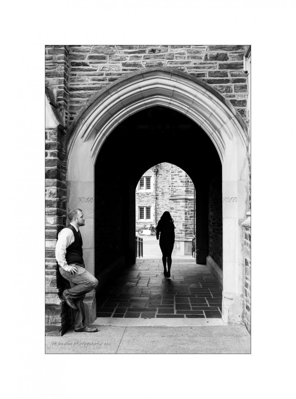 A black-and-white photo of a stone archway. A person leans against the left side, while another walks away through the arch. The intricate stonework and the shadows create a dramatic contrast.