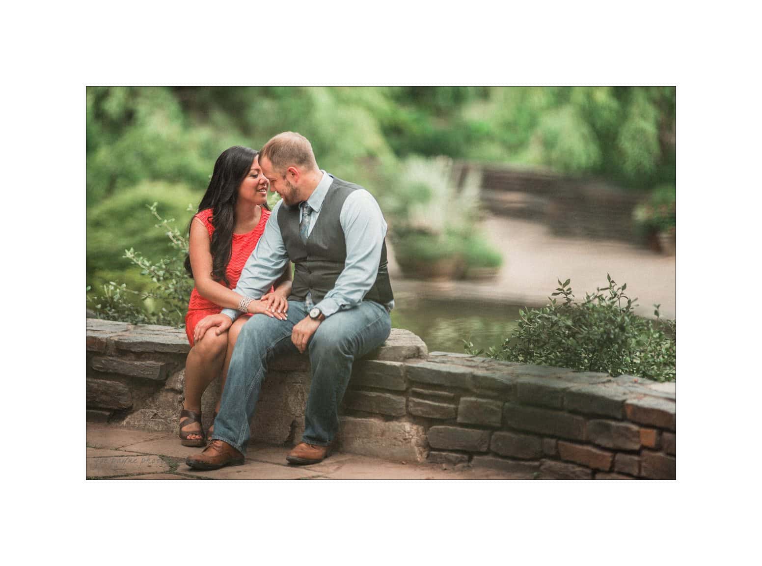 A couple sits on a stone wall, surrounded by lush greenery. The woman is in a red dress, and the man is in a blue shirt with a vest. They are holding hands, smiling at each other, with a serene pond in the background.