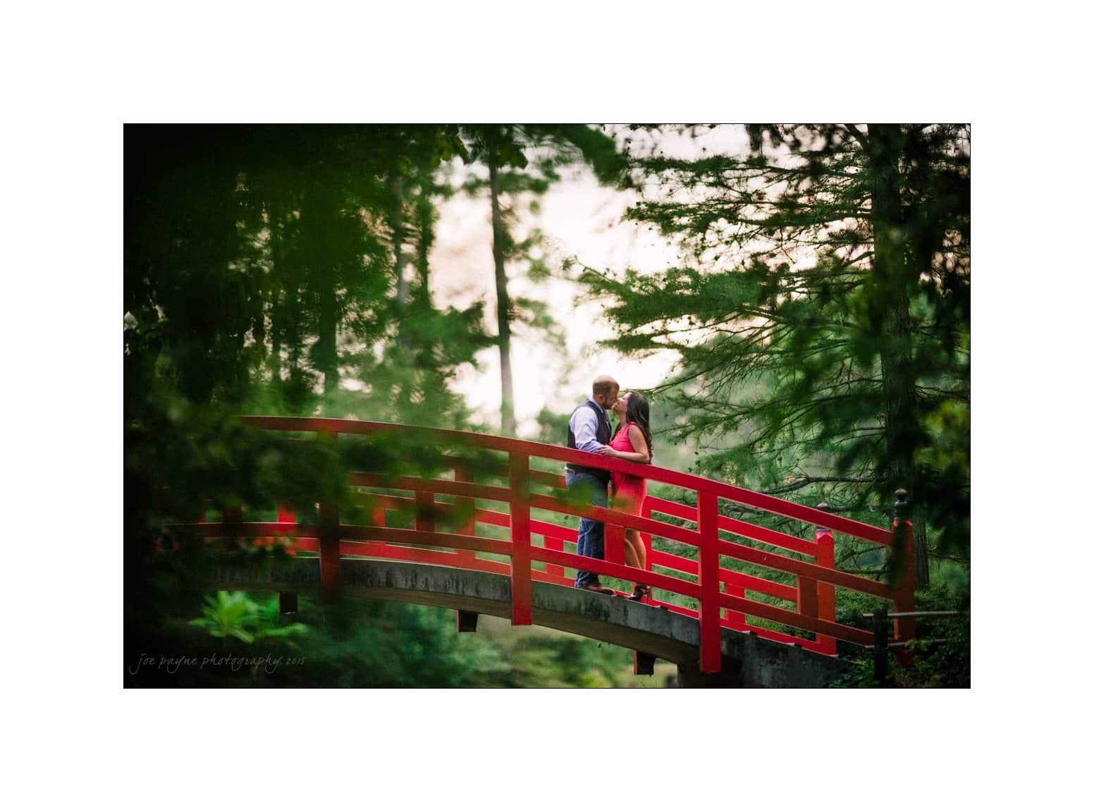 A couple embraces on a red bridge surrounded by lush greenery. The soft focus and vibrant colors create a romantic and serene atmosphere.