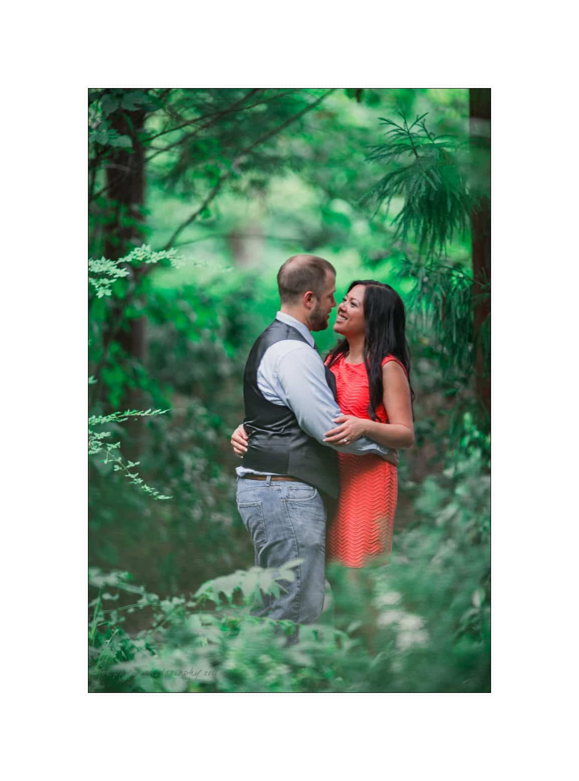 A couple embracing and smiling at each other in a lush, green forest. The man wears a vest and denim jeans, while the woman is in an orange dress. The background is filled with vibrant foliage.