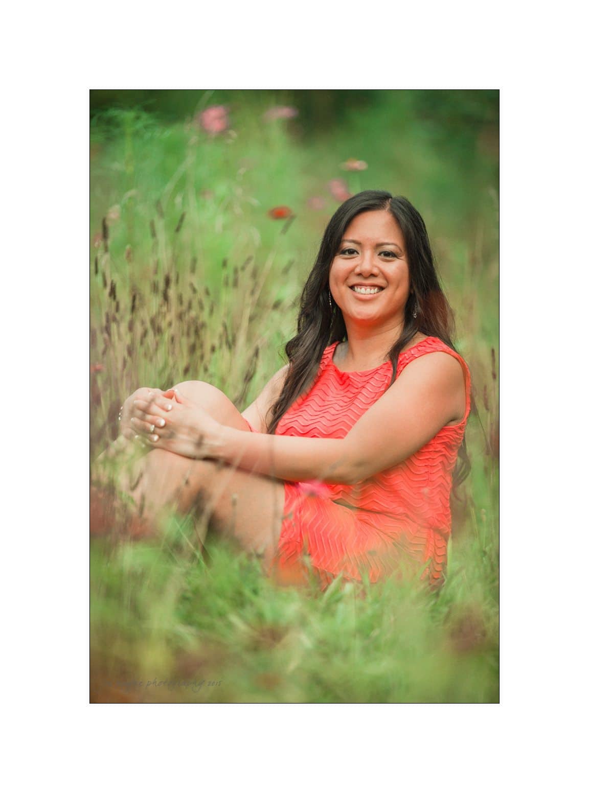 A woman in a vibrant coral dress sits smiling in a lush green field, surrounded by tall grasses and wildflowers. The scene is serene and sunlit, with a soft focus on the natural surroundings.