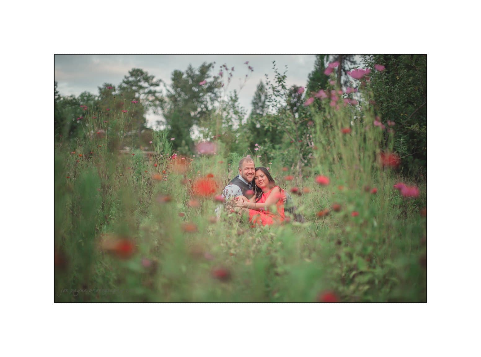 A couple is sitting together in a lush field of wildflowers. They are surrounded by tall green grass and vibrant, blooming flowers. The sky is overcast, creating a soft, serene backdrop.