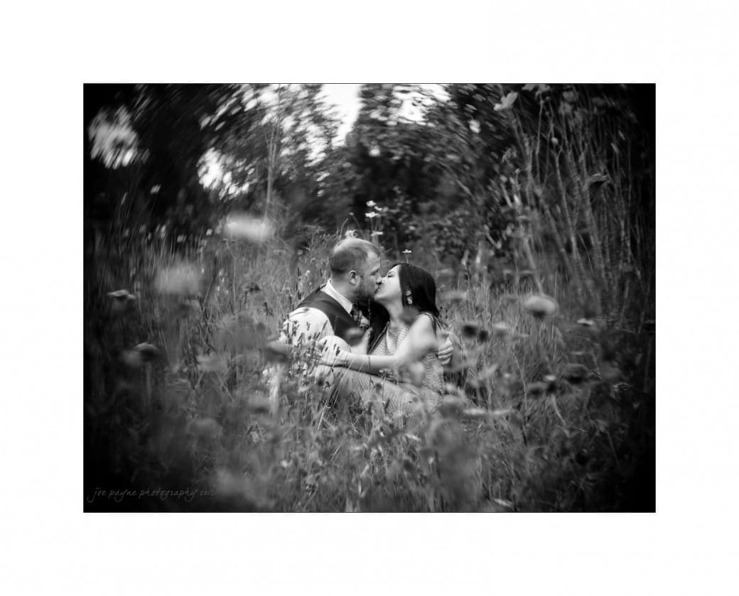 A couple shares a kiss while sitting in a field of tall grass and wildflowers. The image is in black and white with a soft, swirling focus around the edges, creating a dreamy, romantic atmosphere.