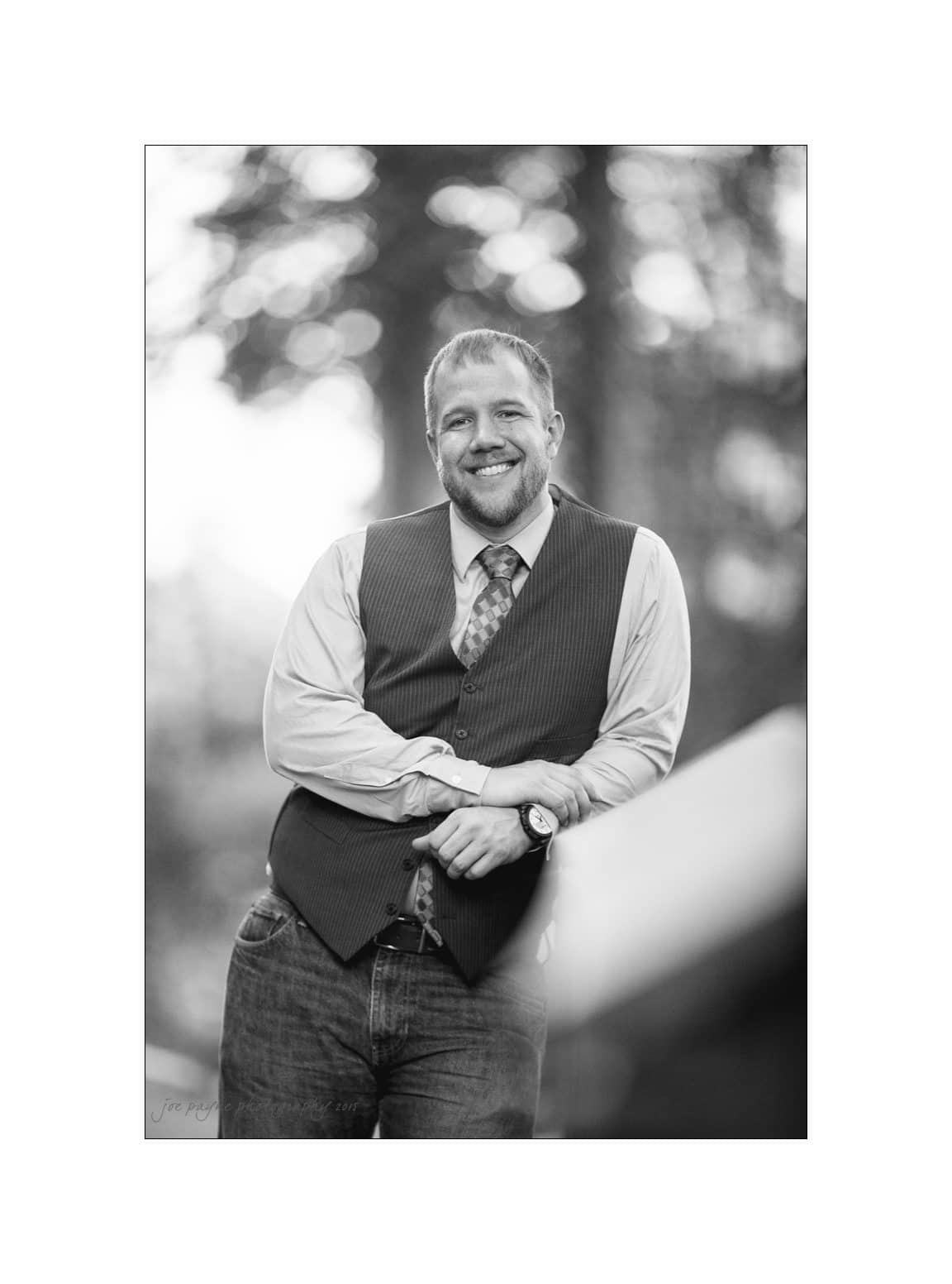 A black-and-white image of a smiling man standing outdoors, wearing a long-sleeve shirt, tie, and vest. He has short hair and facial hair, with one hand resting on the other arm. The background features blurred trees.