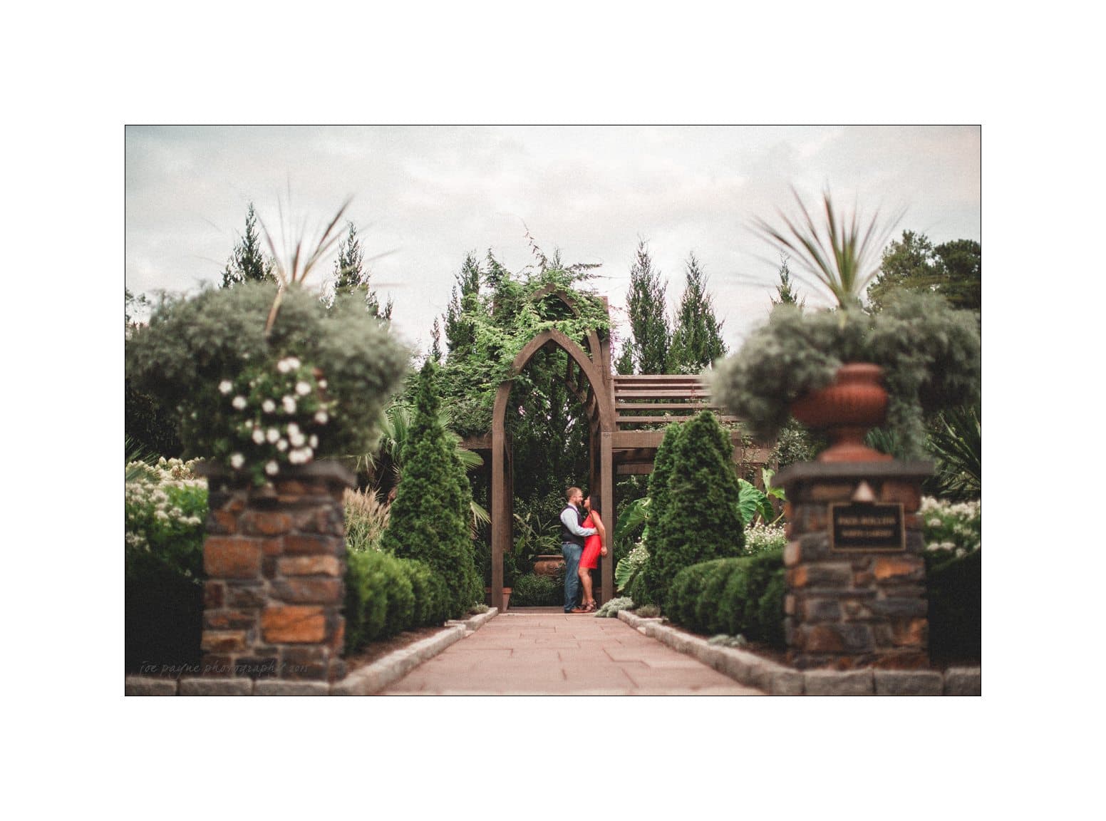 A couple embraces under a wooden archway surrounded by lush greenery and stone pillars in a serene garden setting. The sky is overcast, adding a soft light to the romantic scene.