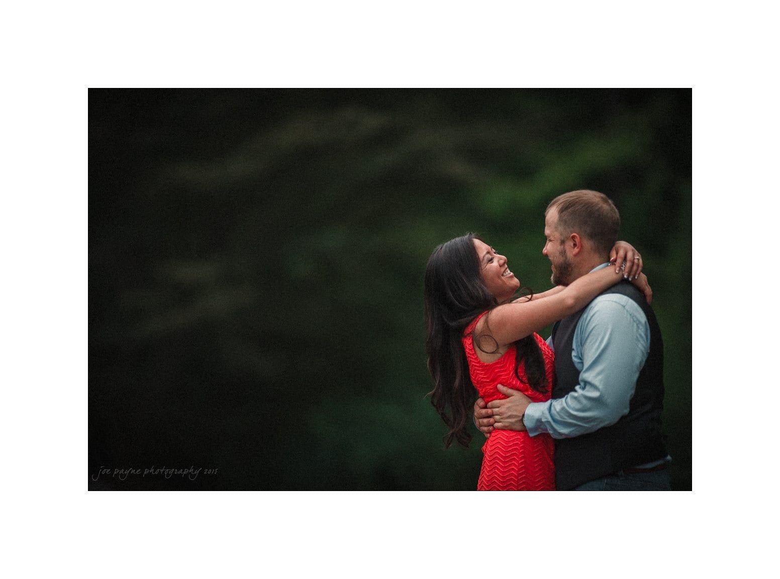 A couple embraces joyfully in an outdoor setting. The woman in a red dress has her arms around the mans neck, while he holds her waist. Both are smiling warmly at each other, against a blurred natural background.