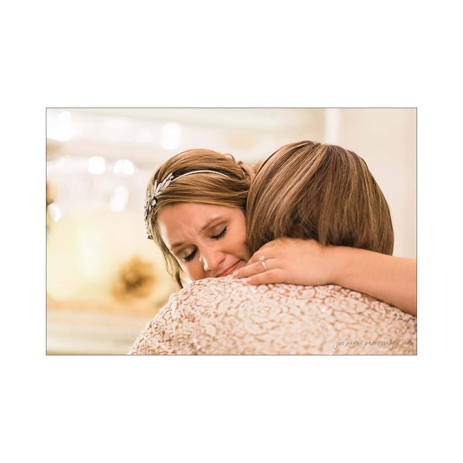 A bride with closed eyes and a tiara hugs another person closely, showing emotion. The background is softly blurred, suggesting a sentimental moment at a wedding.