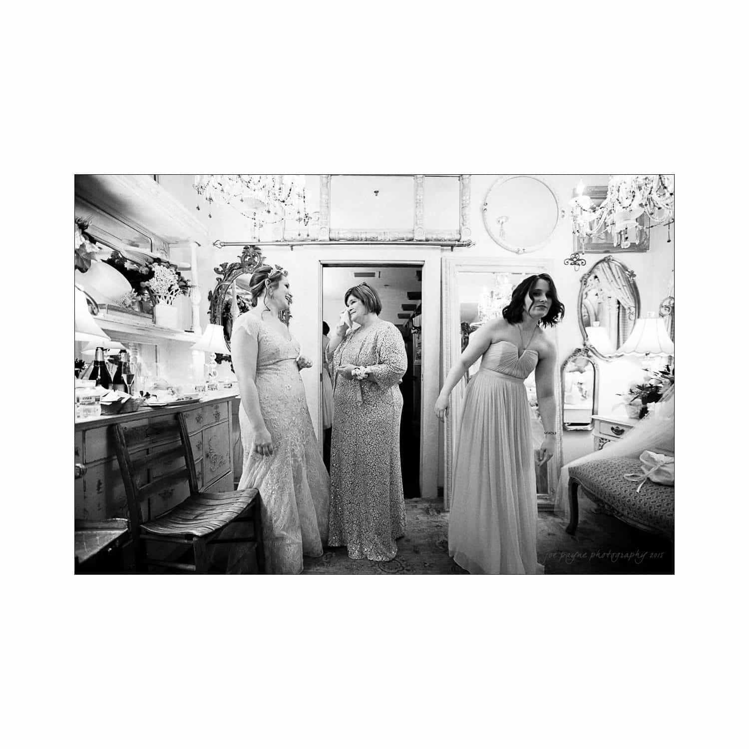A black and white photo of four women dressed in elegant gowns, standing in a vintage-styled room with mirrors, chandeliers, and floral arrangements. They appear to be laughing and preparing for a special occasion.