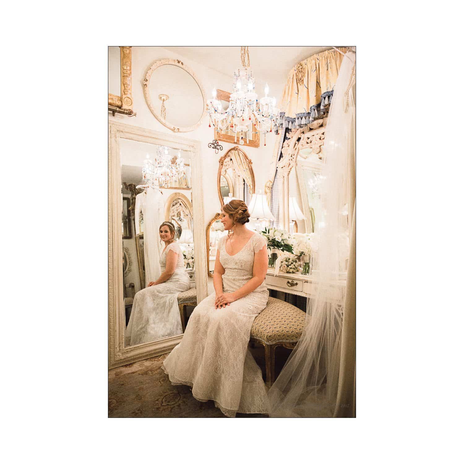 A bride in an elegant white dress sits on a cushioned bench in a luxurious room. She is looking into a large mirror, surrounded by ornate decor, chandeliers, and floral arrangements. The ambiance is romantic and vintage.