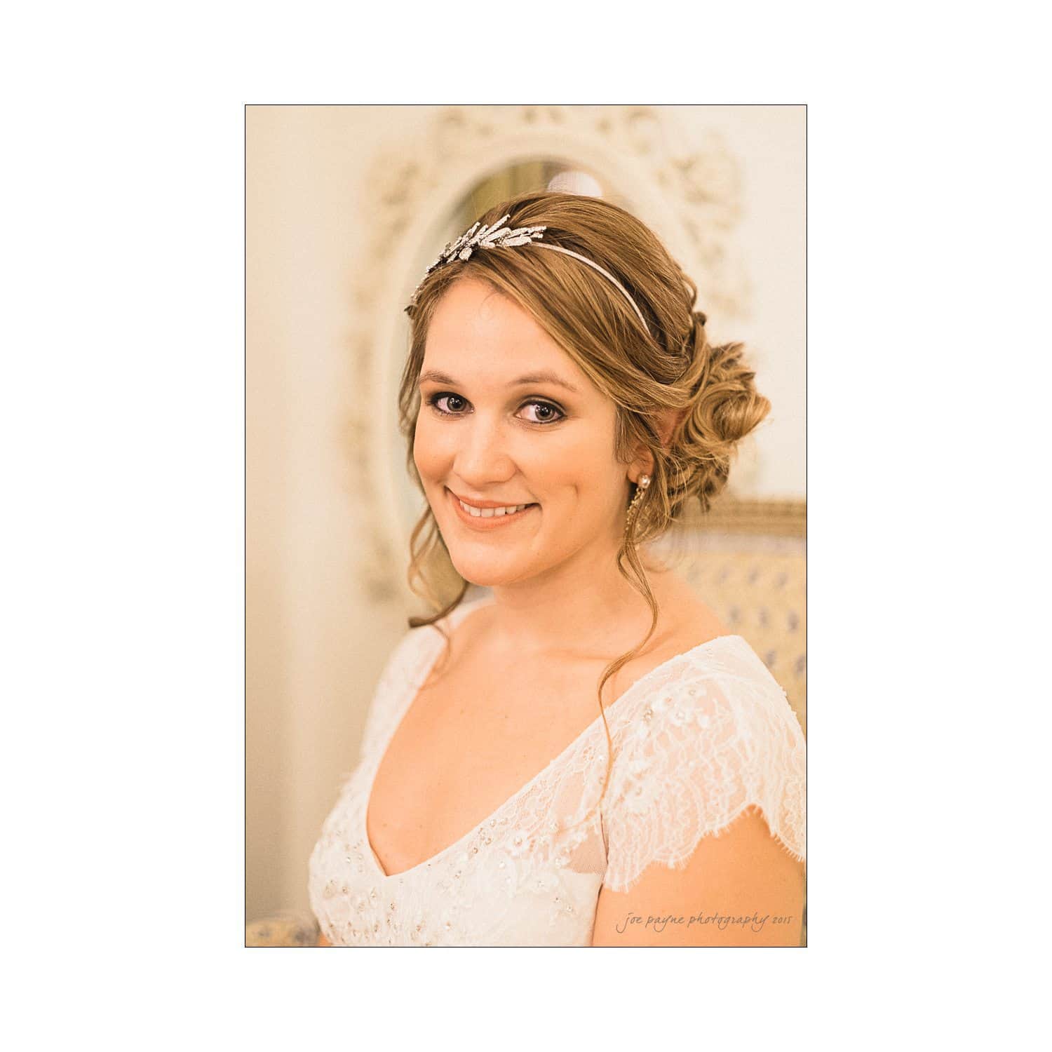 A bride smiling, wearing a white lace wedding dress and a delicate headband. Her hair is styled in an elegant updo. She is seated, with a soft, ornate background visible.