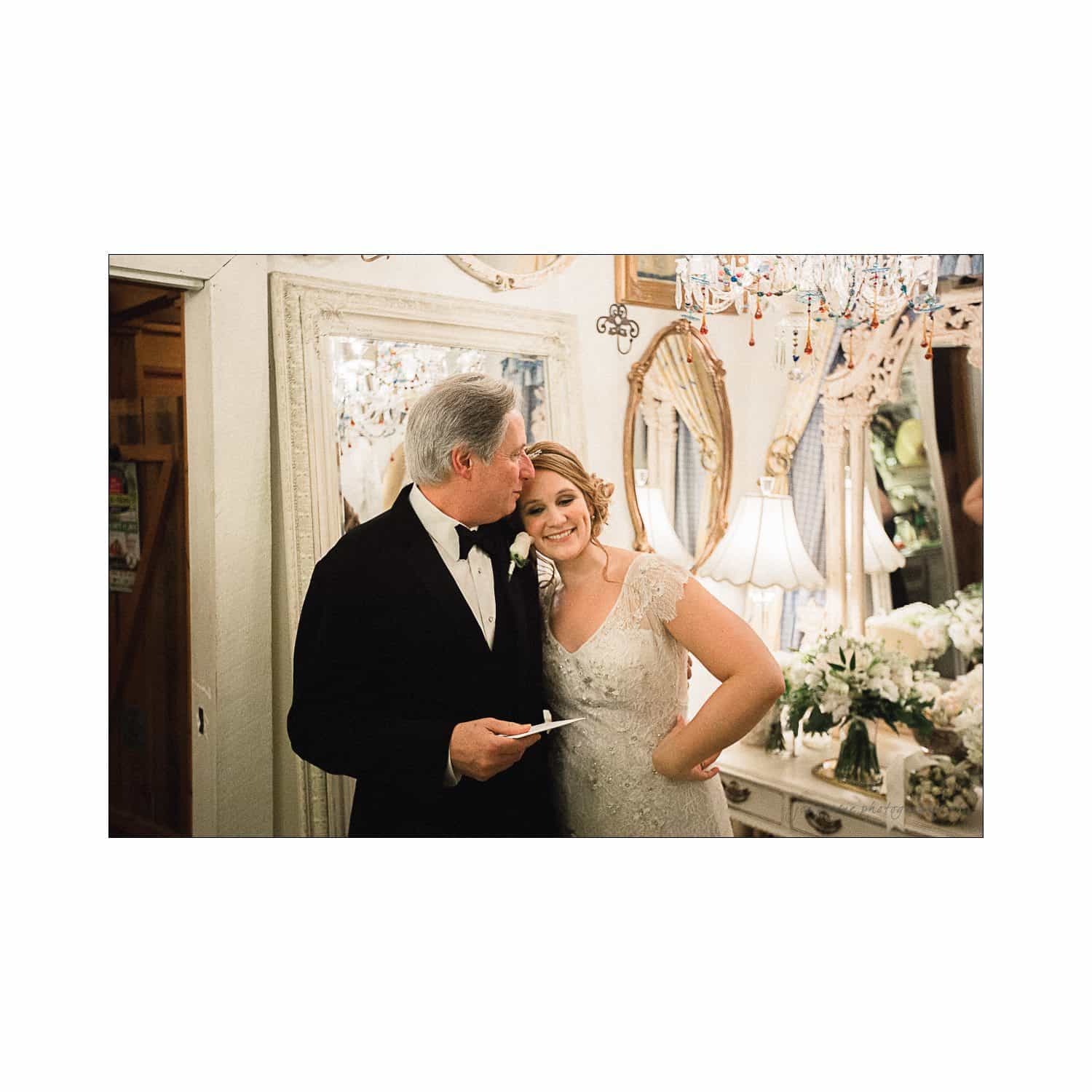 A man in a tuxedo kisses the forehead of a smiling woman in a white dress, standing close together inside a room adorned with white flowers, mirrors, and a chandelier.