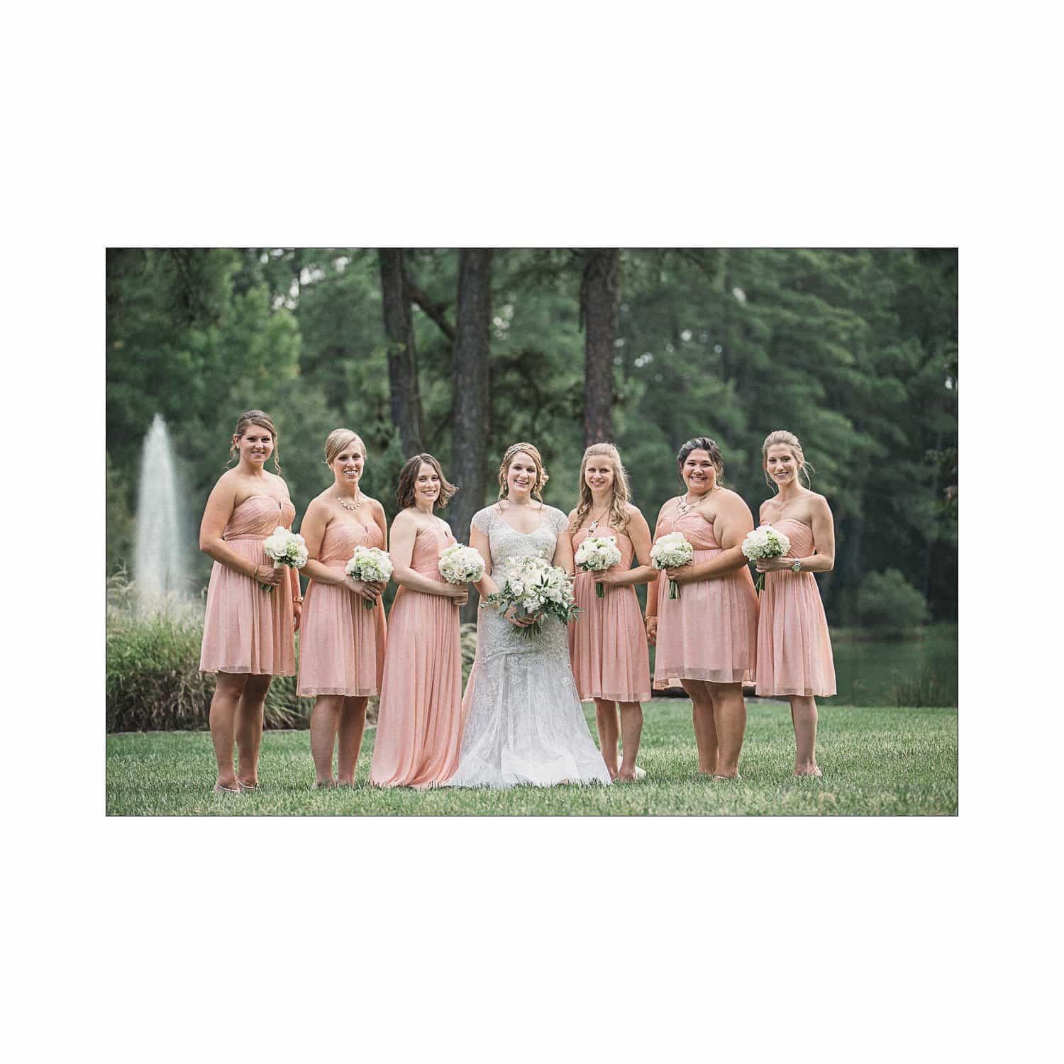 A bride stands with six bridesmaids in a park. The bride wears a white gown, and the bridesmaids wear matching pale pink dresses. They all hold white bouquets. Trees and a fountain are visible in the background.