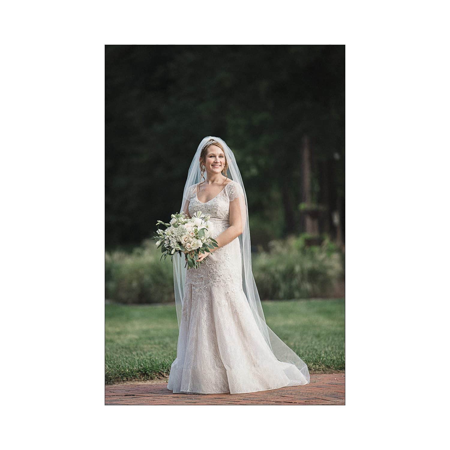 A bride in a lace wedding dress stands outdoors, holding a bouquet of white flowers. She smiles, with a long veil flowing behind her. The background features greenery and a blurred garden setting.