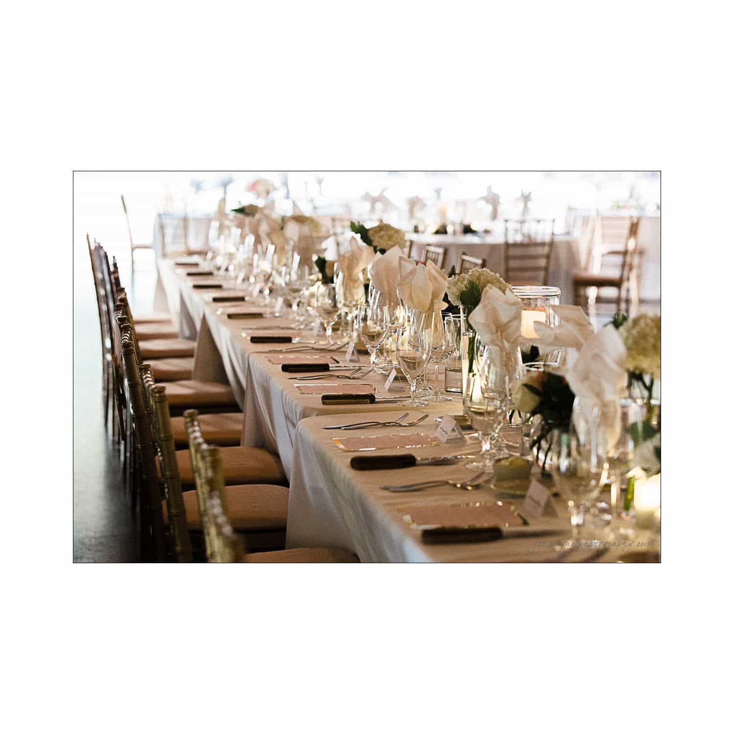 An elegantly set dining table with white linens, white napkins, and crystal glassware. Soft lighting enhances the ambiance, with floral centerpieces along the table.