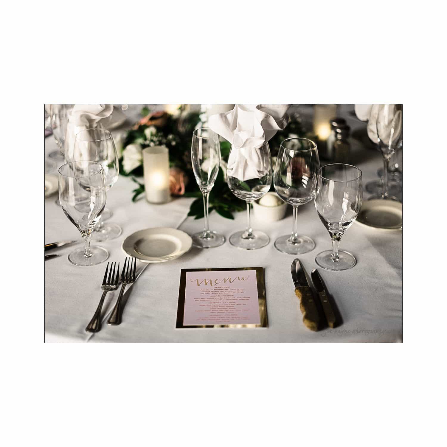 A beautifully set wedding table features wine glasses, folded white napkins, silverware, a candle, and a floral centerpiece. A pink menu lies prominently on the white tablecloth.