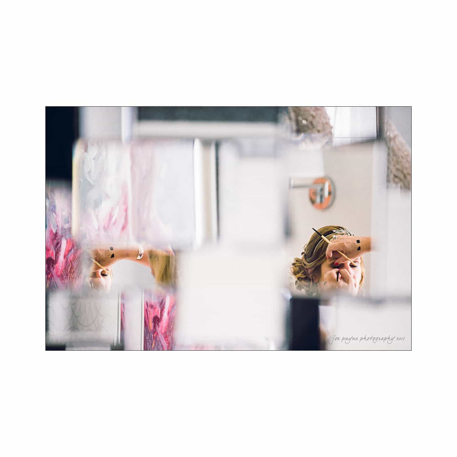 A womans face is partially visible through fragmented, reflective surfaces creating an abstract, mirrored effect. She appears to be looking up or adjusting something above her. The background is blurred with hints of color.