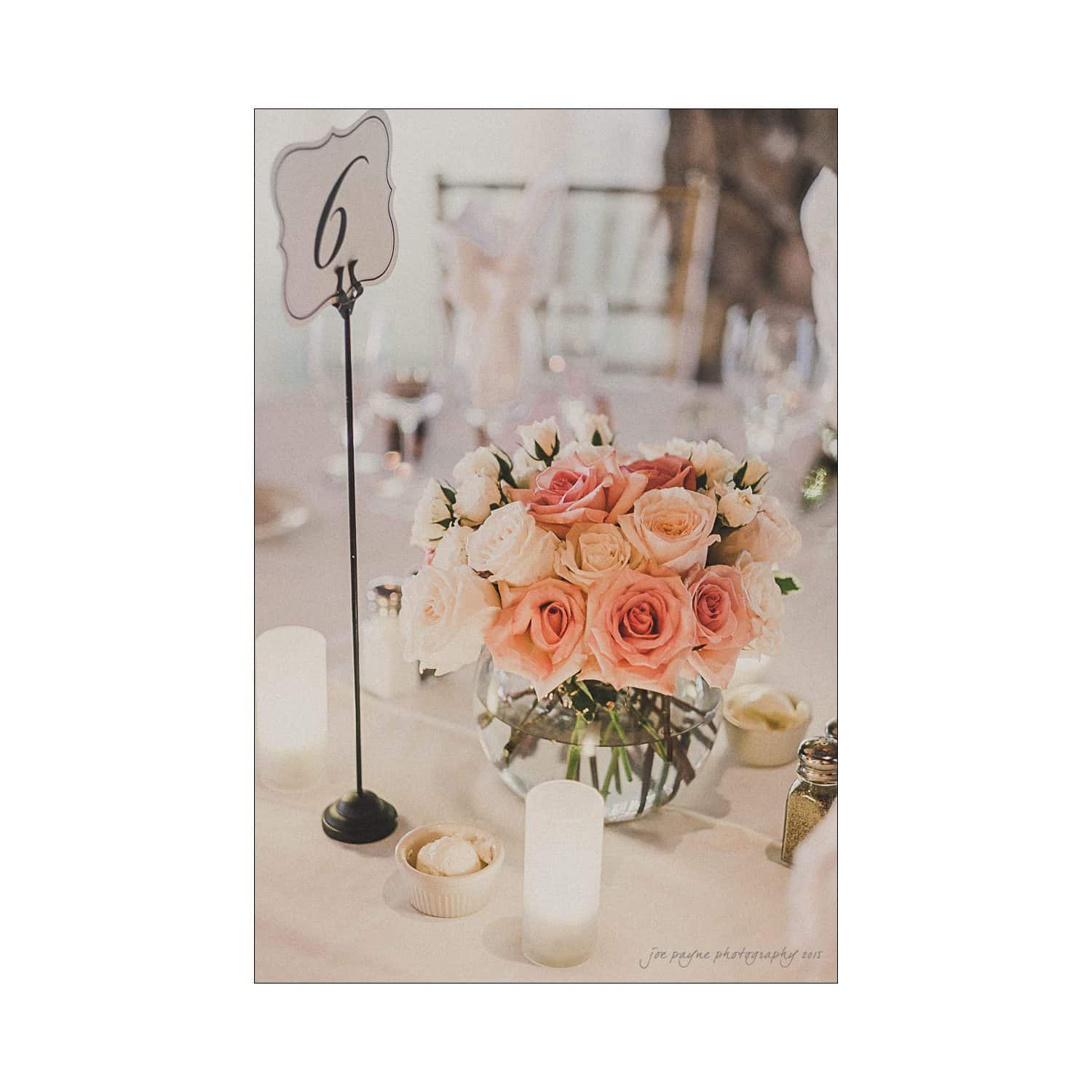 A wedding table centerpiece features a round vase filled with pink and white roses. The table is decorated with candles, a small dessert, and a table number 6 sign on a stand.