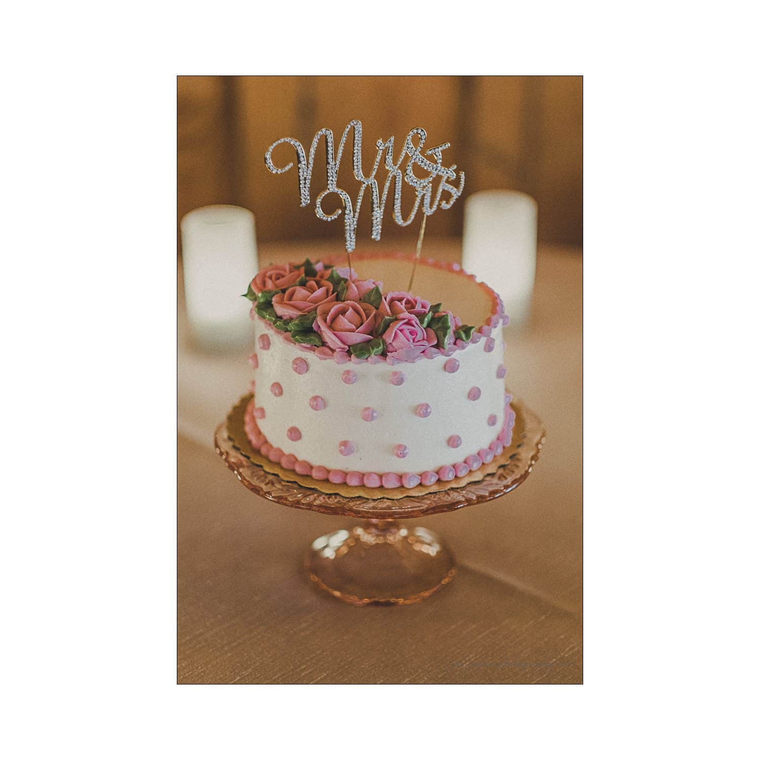 A white cake decorated with pink frosting dots and pink roses on top, placed on a gold cake stand. The cake is topped with a Mrs & Mrs sign, with two candles in the background.