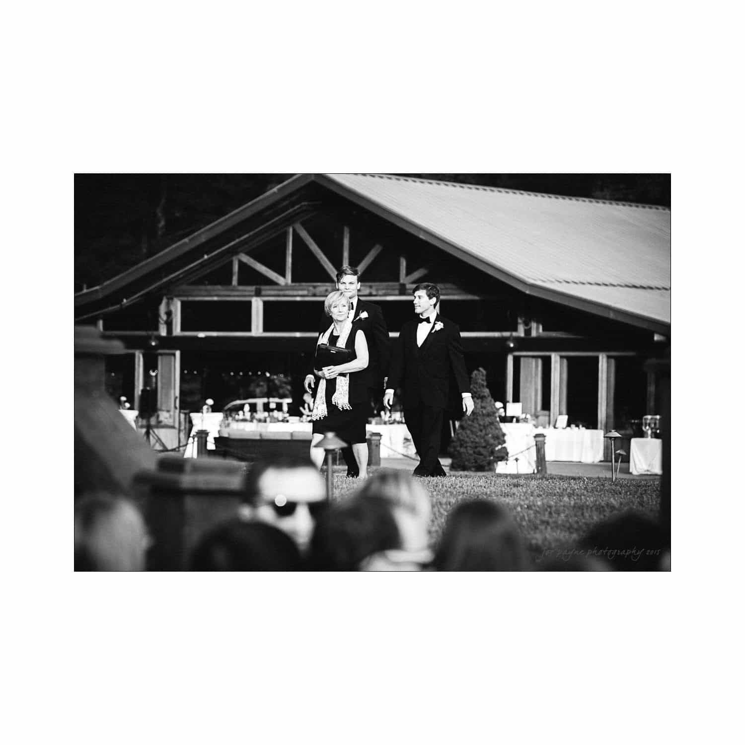 A black-and-white photo of a formal event. A man and woman in elegant attire walk outdoors in front of a building with wooden beams. The background features tables and a large tree.