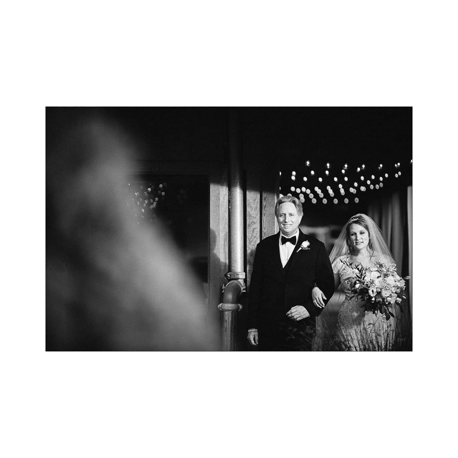 A black and white photo of a bride walking arm in arm with a man in a tuxedo. The bride holds a bouquet and wears a veil. String lights are visible in the background, creating a festive atmosphere.