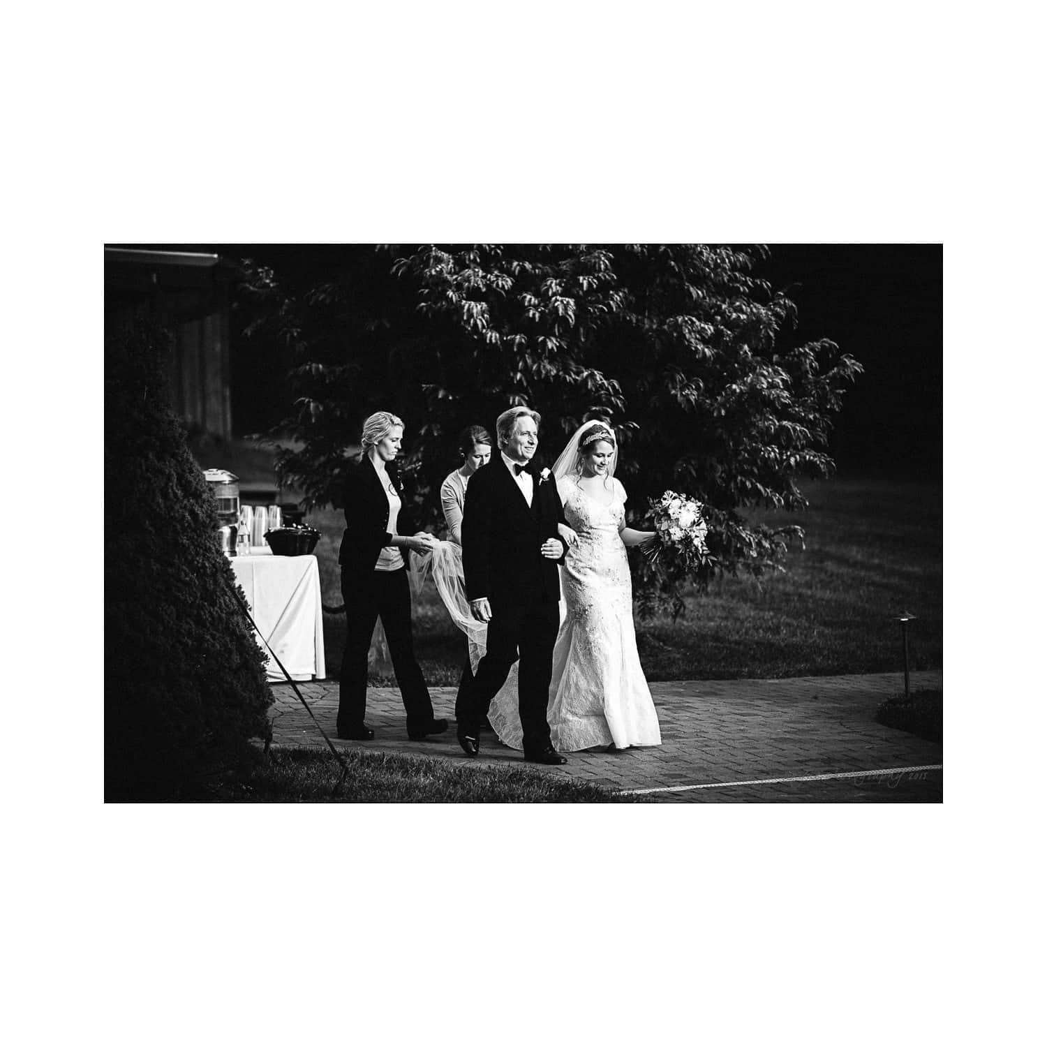 A bride and groom walk arm in arm on a paved path, surrounded by trees and grass. The bride wears a long white dress and holds a bouquet. Another person follows behind them, with a table and canopy visible in the background.