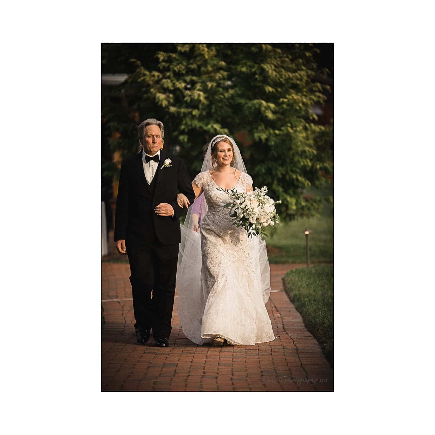 A bride in a white wedding dress, holding a bouquet, walks down an outdoor aisle alongside a man in a tuxedo. Lush greenery surrounds the path, creating a serene, romantic atmosphere.