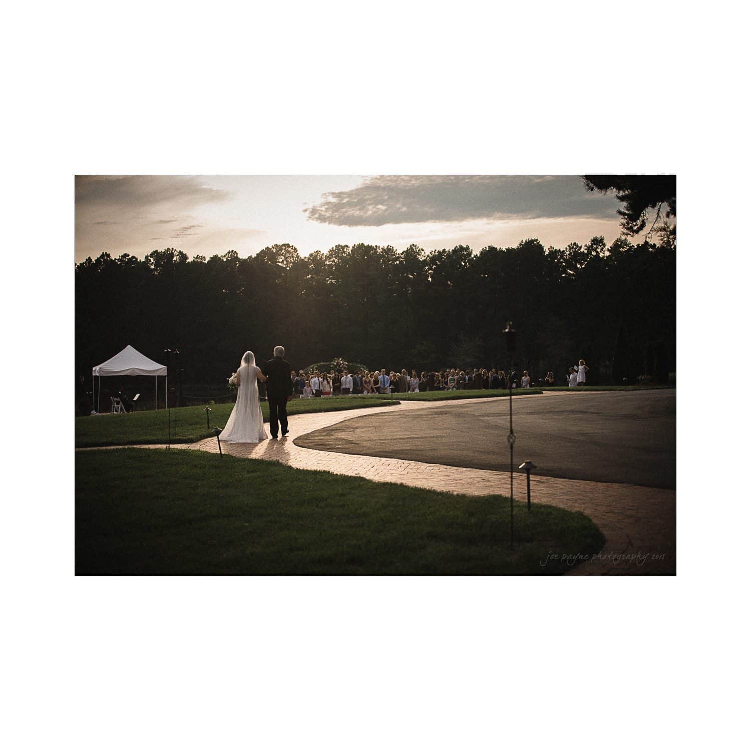 Bride and father walking down an outdoor aisle at sunset, with guests seated in the background surrounded by trees and a white tent nearby.